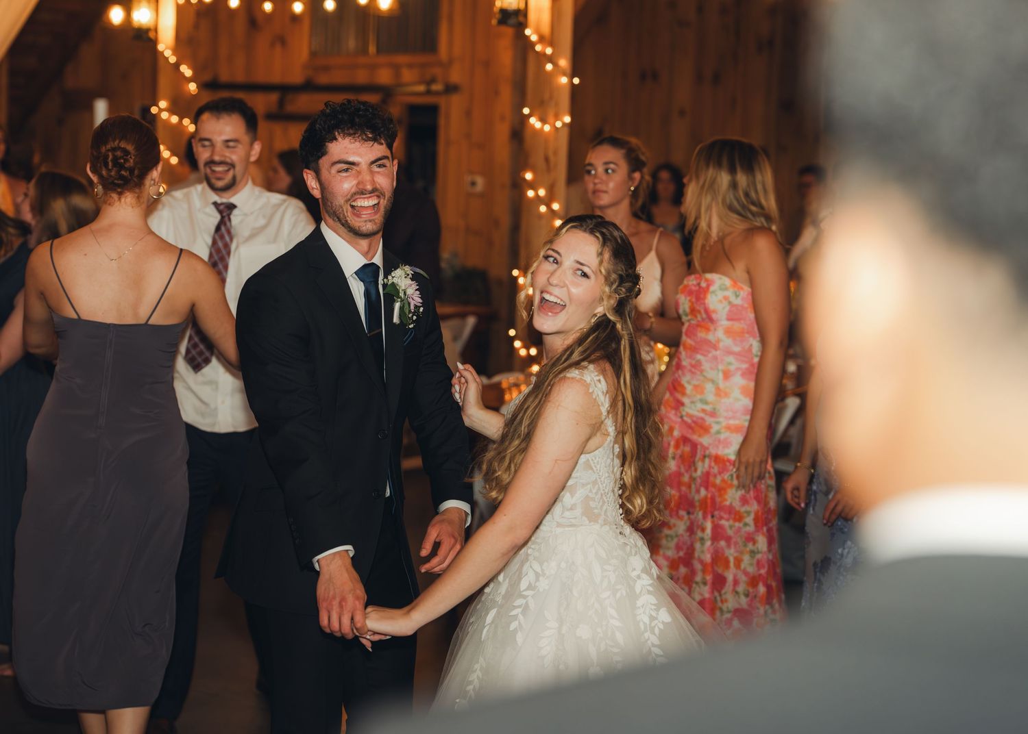 Bride and groom laughing and dancing joyfully with guests on the dance floor at their rustic barn reception.