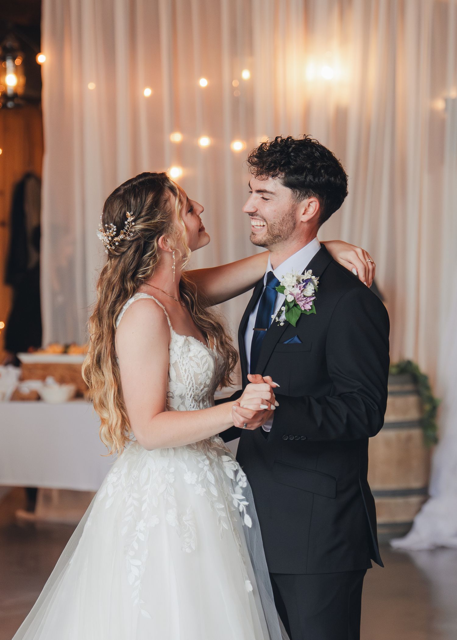 Bride and groom share their first dance, smiling at each other in elegant wedding attire with fairy lights backdrop.