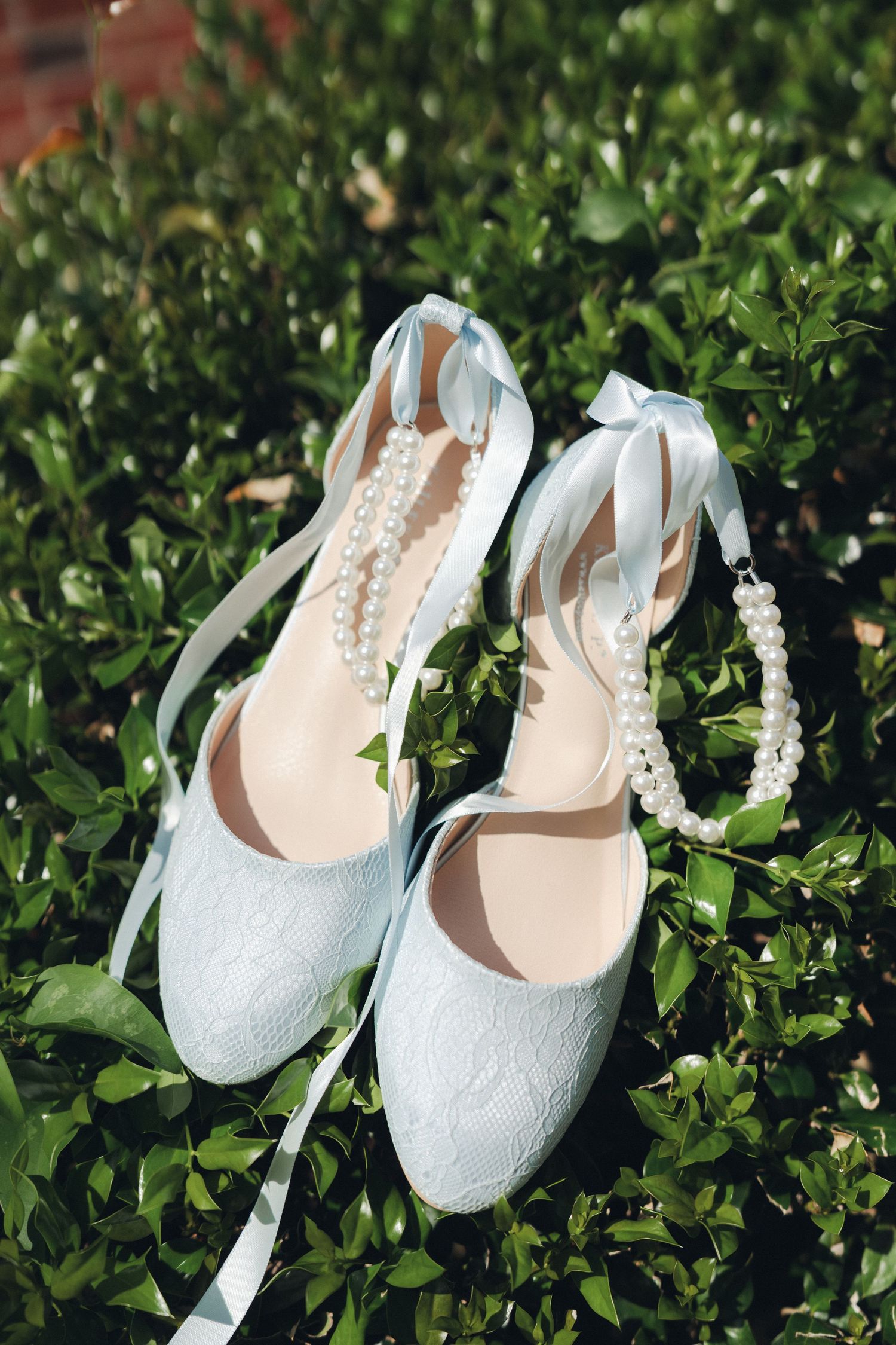 Light blue lace bridal heels with satin ribbons and pearl necklace displayed on green garden foliage.