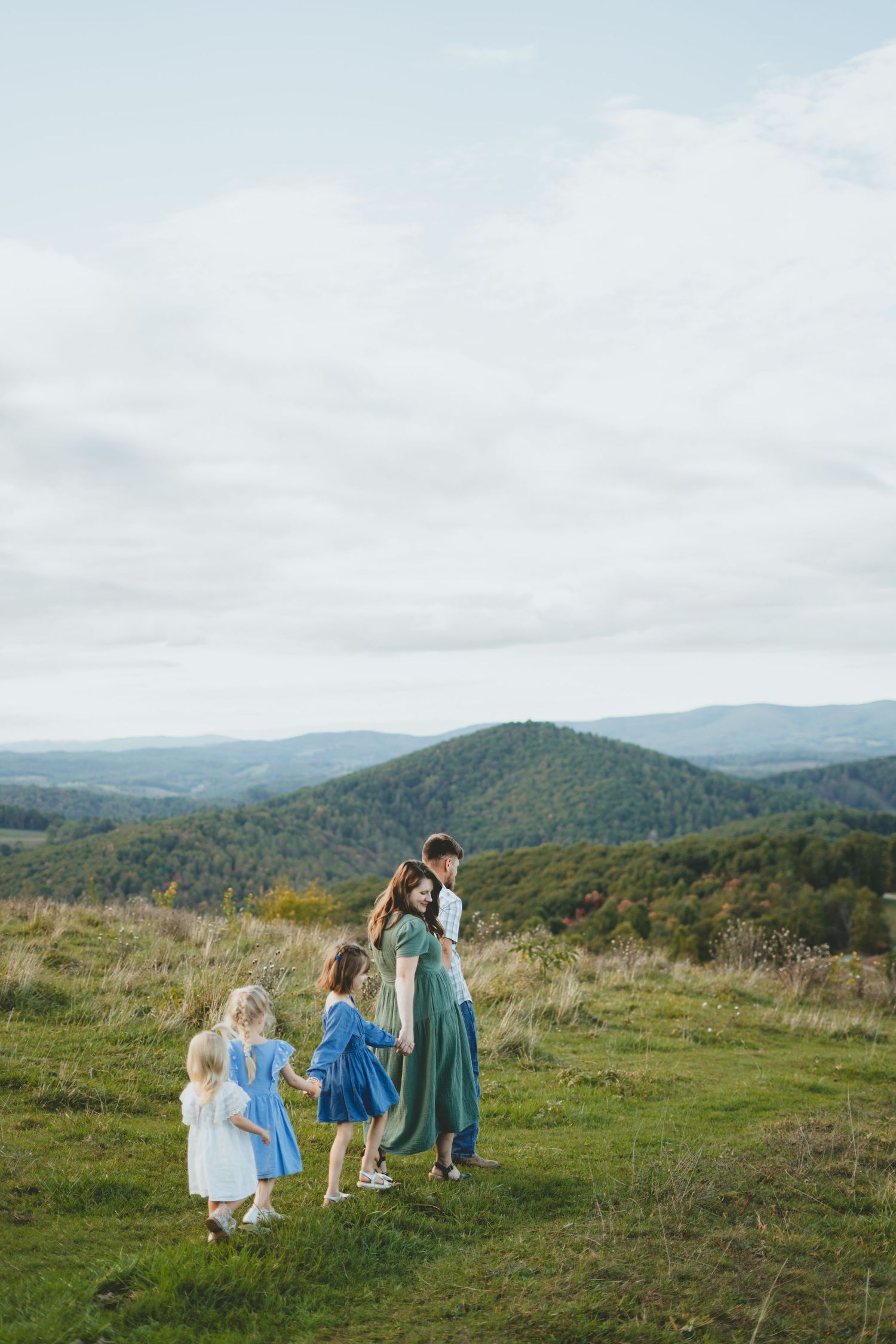Family with three young girls in blue dresses standing on a scenic mountain overlook with rolling hills.