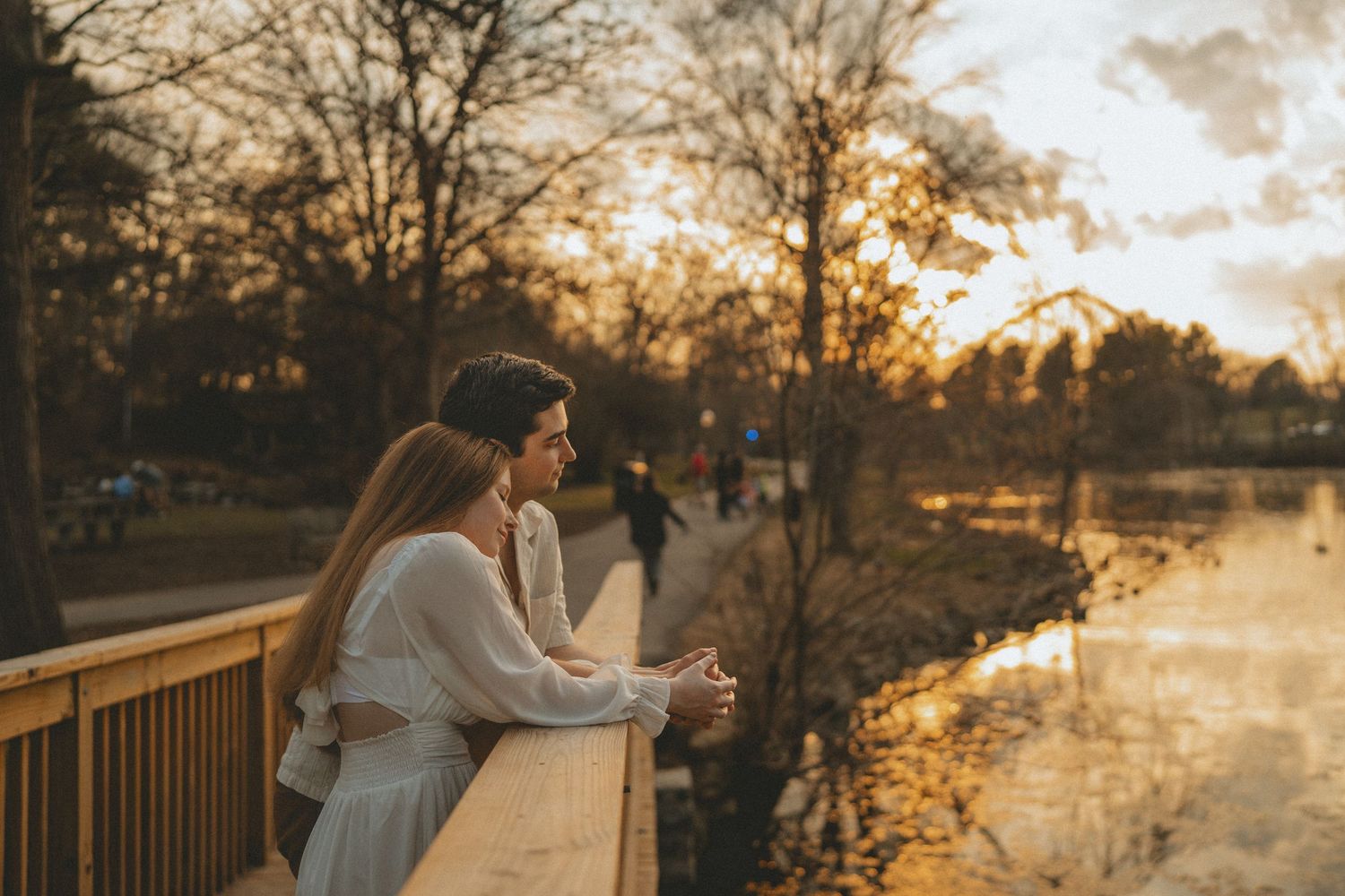 Romantic couple leaning on wooden bridge railing watching sunset reflection over calm park lake.