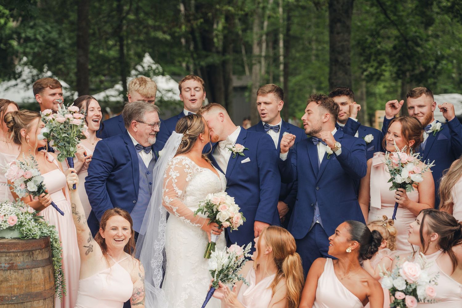 Wedding party celebrating with bride and groom kissing, bridesmaids in blush pink and groomsmen in navy blue suits outdoors.