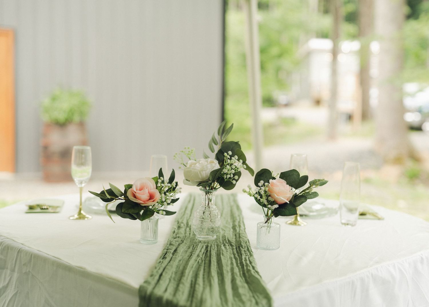 Elegant wedding sweetheart table with green runner, pink roses, and greenery centerpieces on white linen.