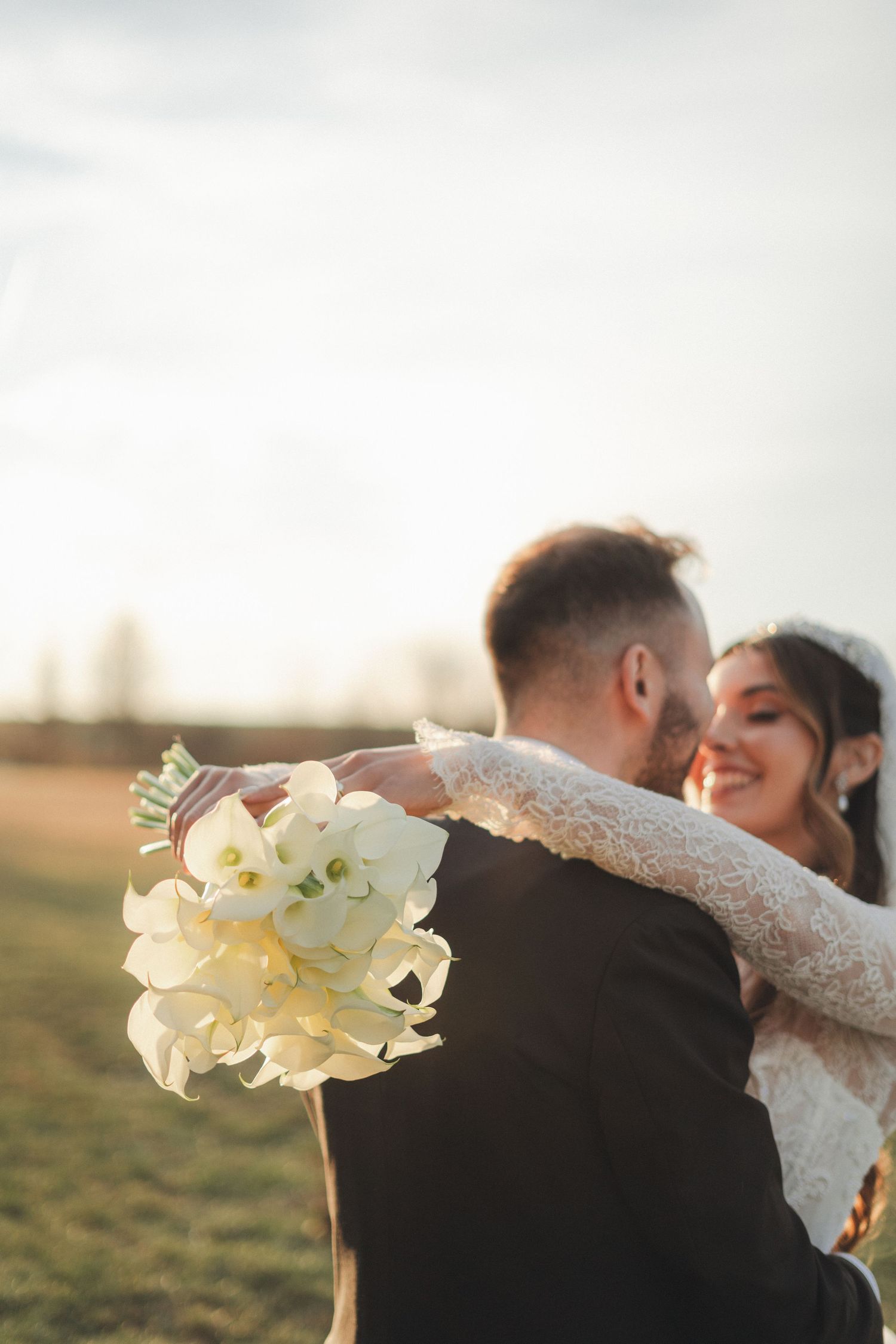 Smiling bride embracing groom outdoors at sunset while holding white floral wedding bouquet.
