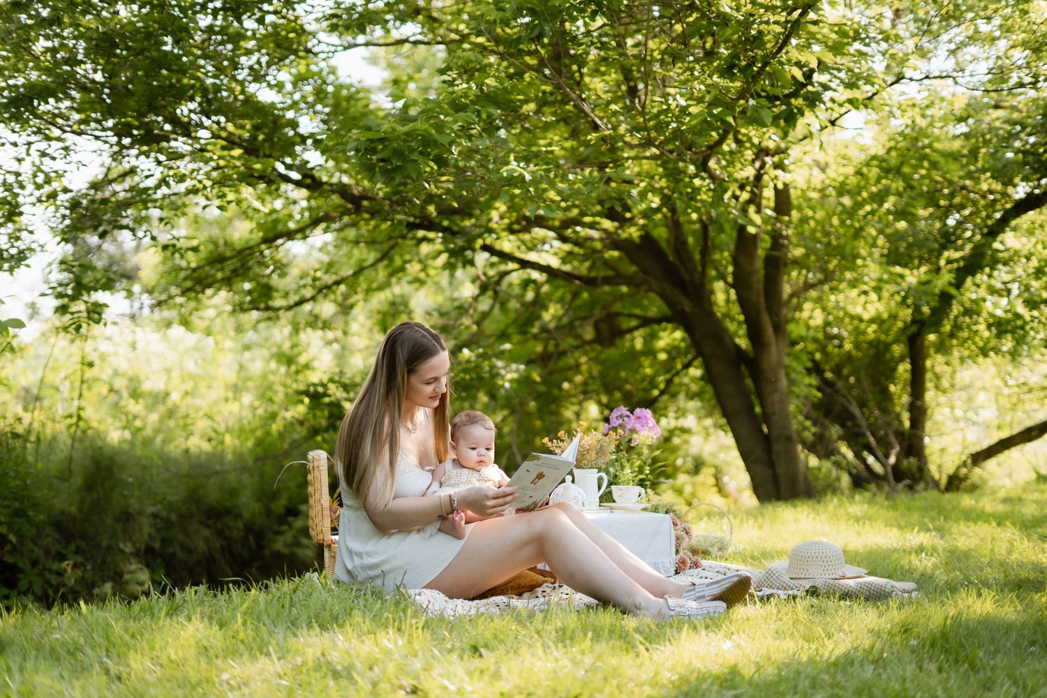 Young mother reading a book with her toddler on a blanket under a shady green tree.
