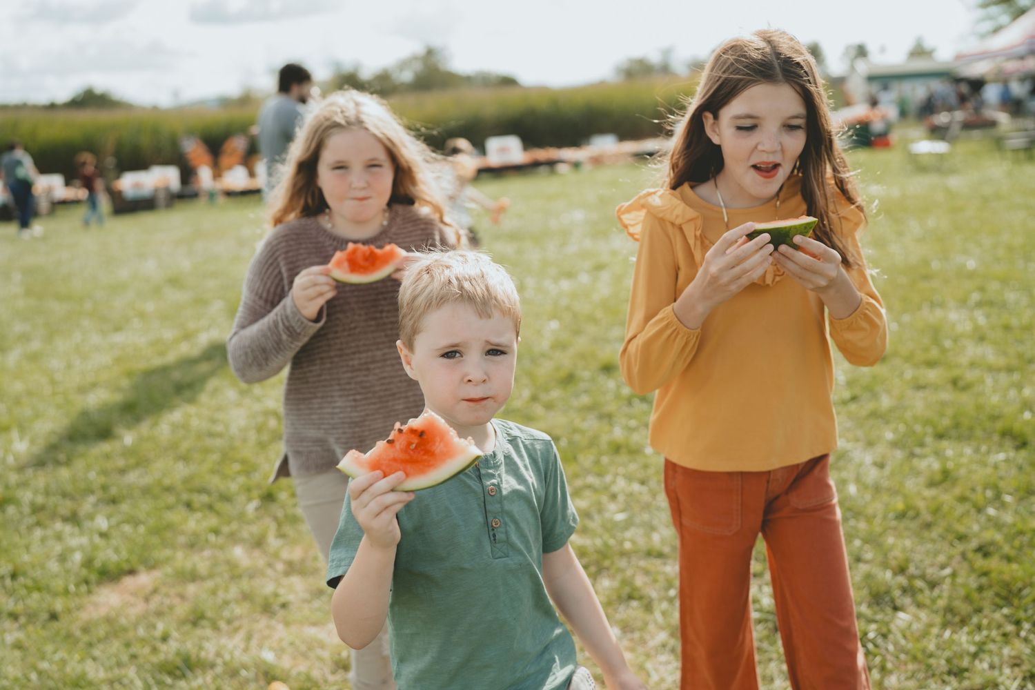 Three children eating watermelon slices outdoors in a grassy field on a sunny day.