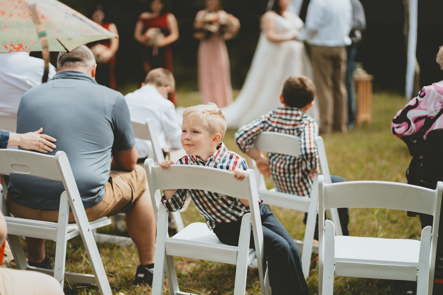 Young blond boy sitting on white folding chair turning around during an outdoor wedding ceremony.