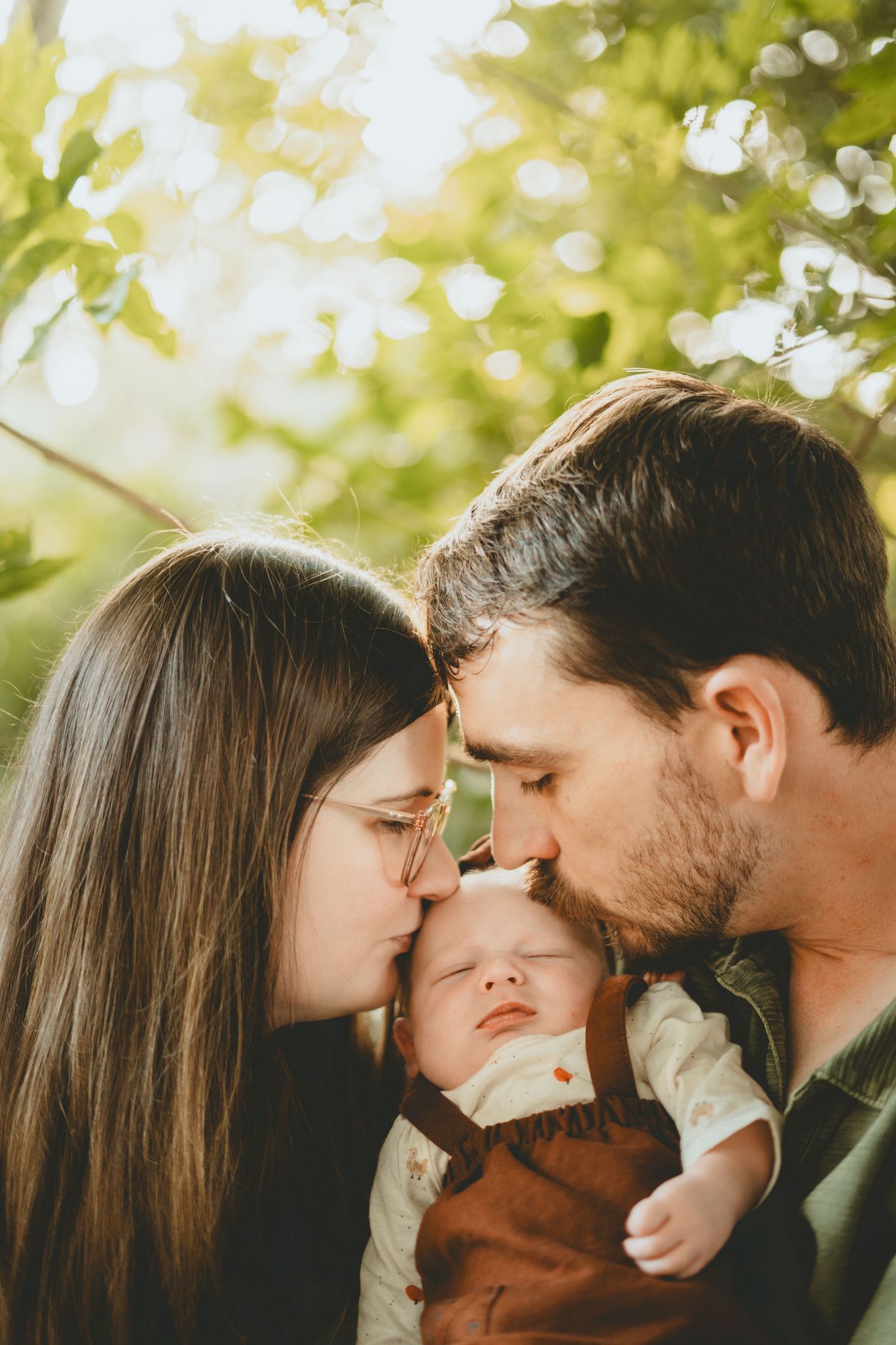 Parents tenderly kiss their newborn baby outdoors in golden sunlight among green foliage.