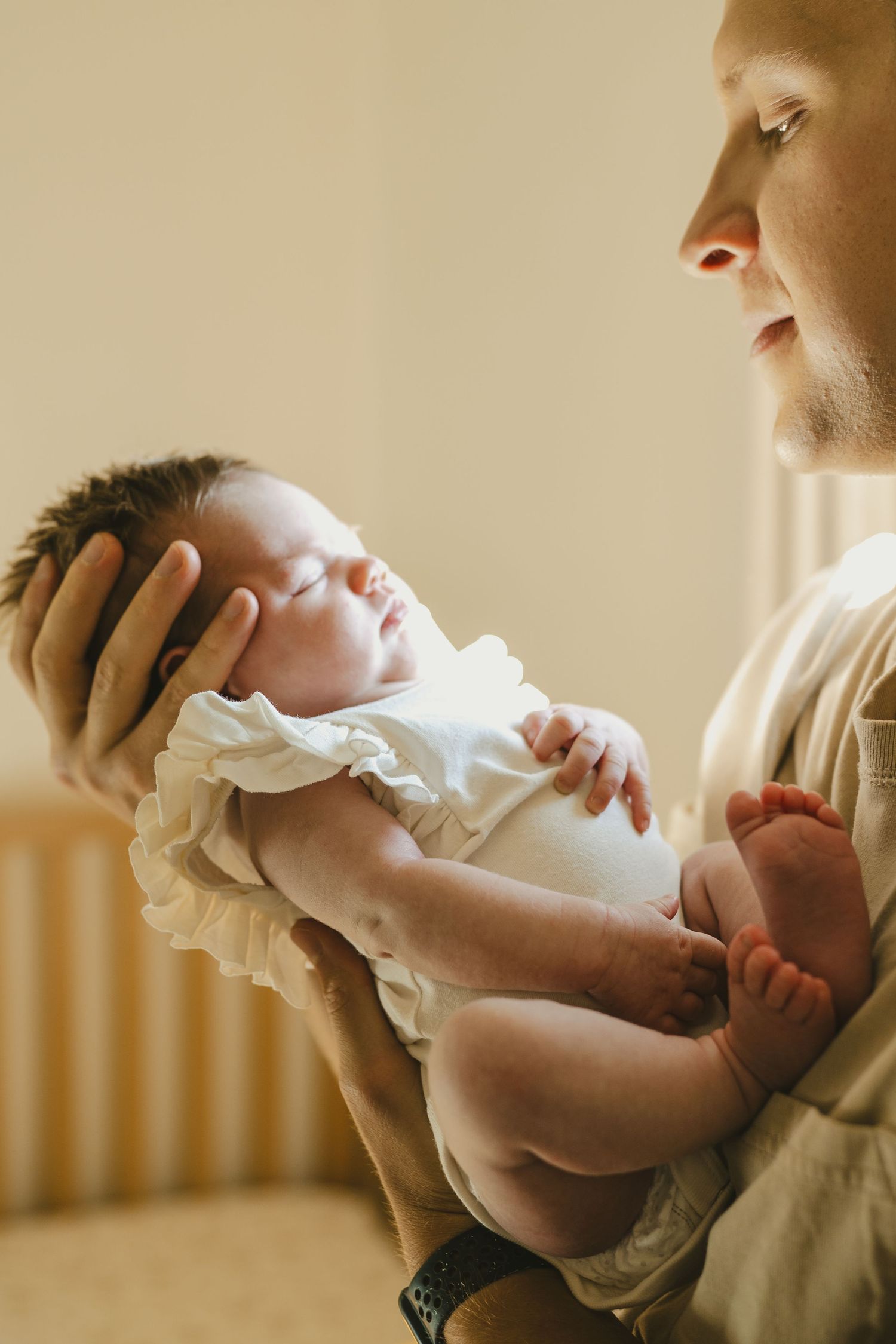 Father tenderly holding sleeping newborn baby girl in white ruffle onesie in warm nursery light.