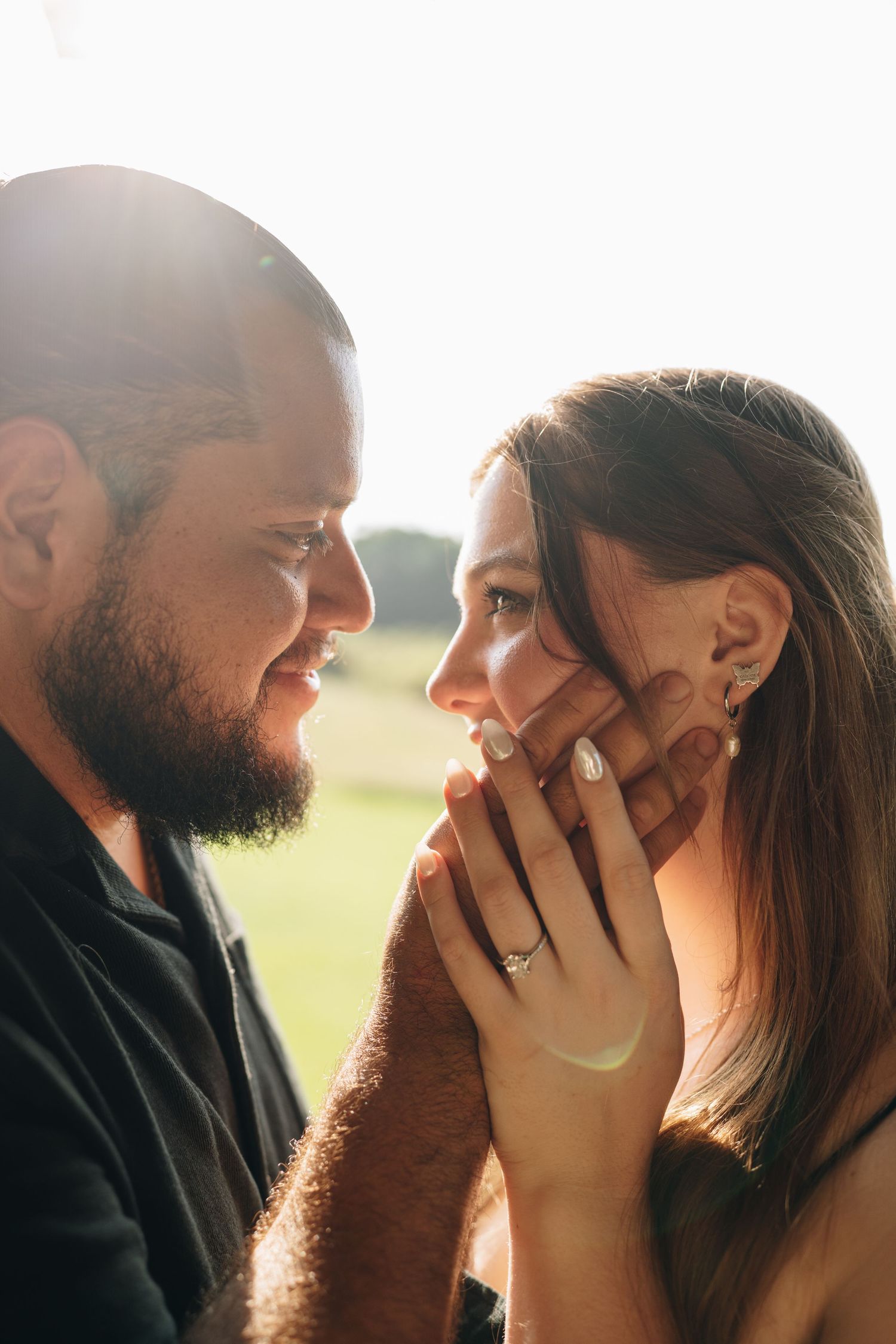 Bearded man smiling at woman holding her face with engagement ring visible in sunlight.