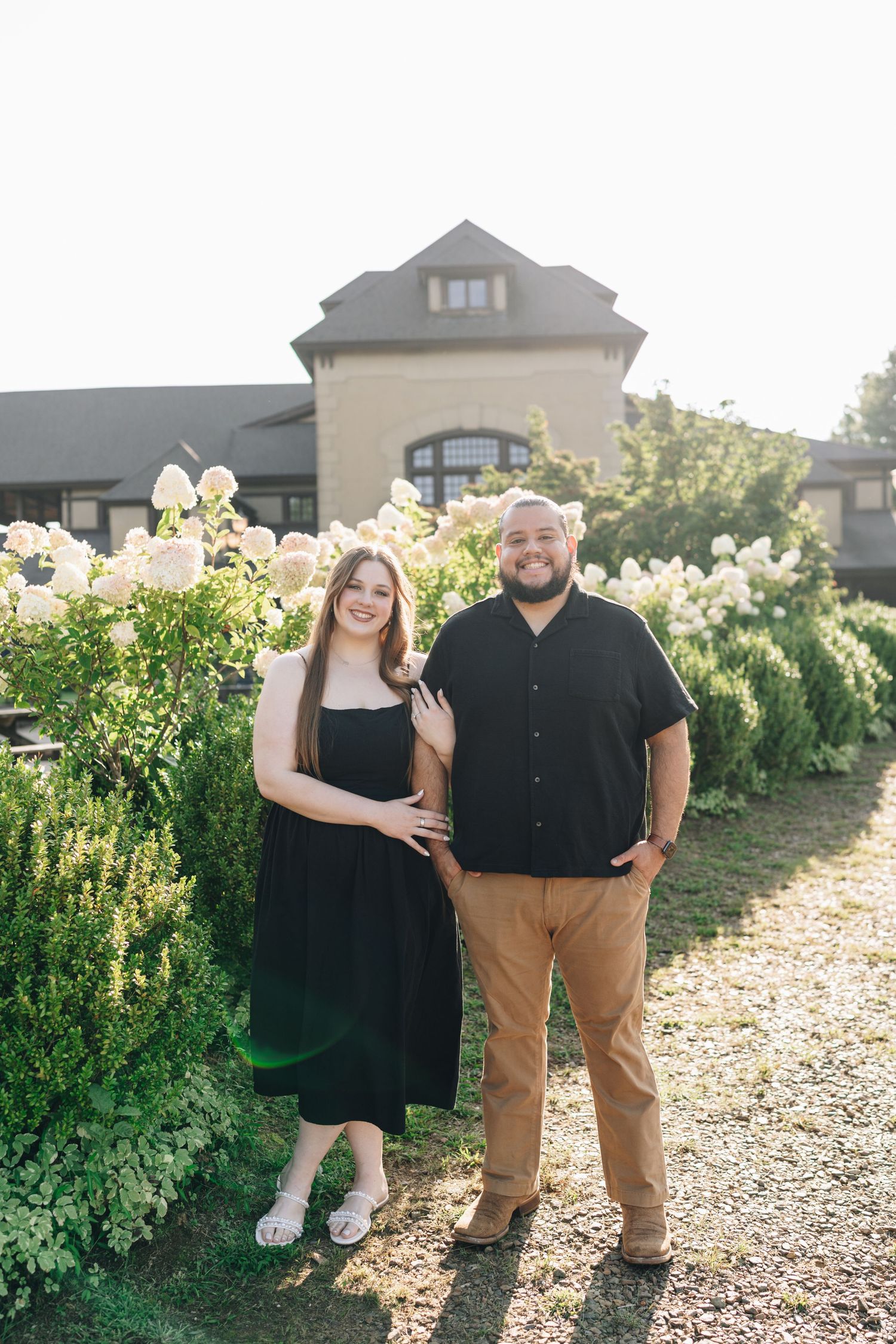 Smiling couple posing together beside lush white hydrangea bushes in front of a beautiful estate.
