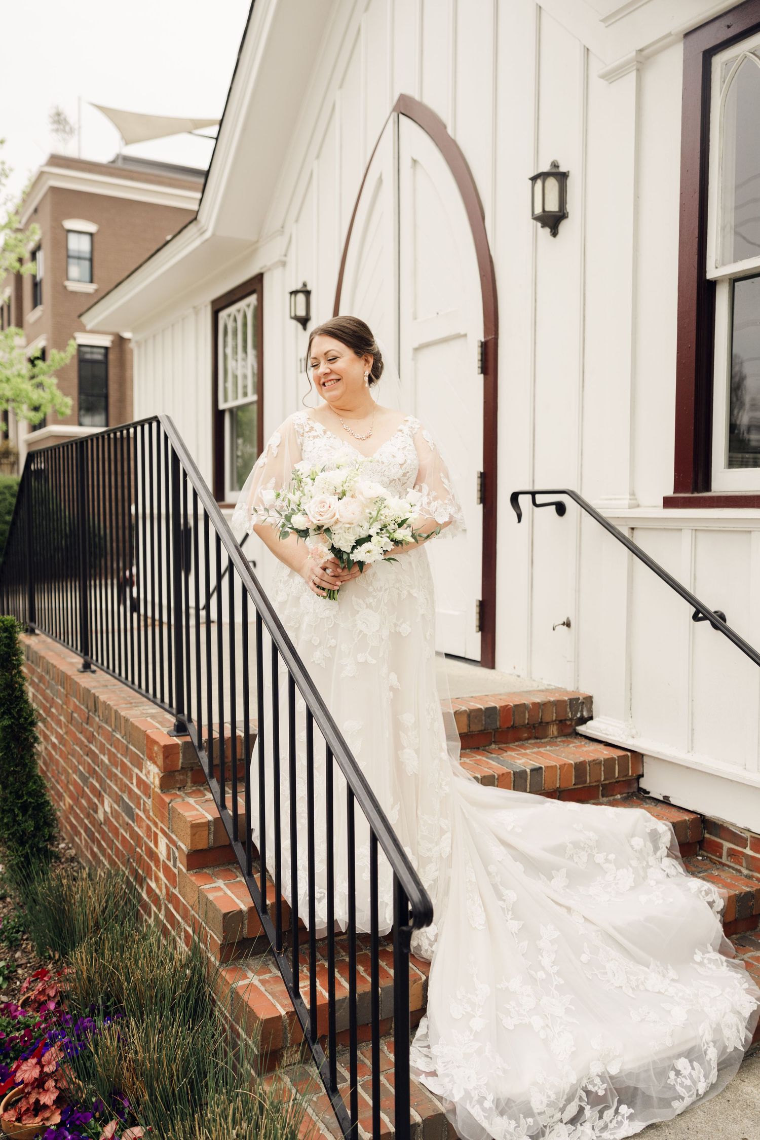 Smiling bride in lace wedding dress holds white bouquet on brick steps outside white chapel.