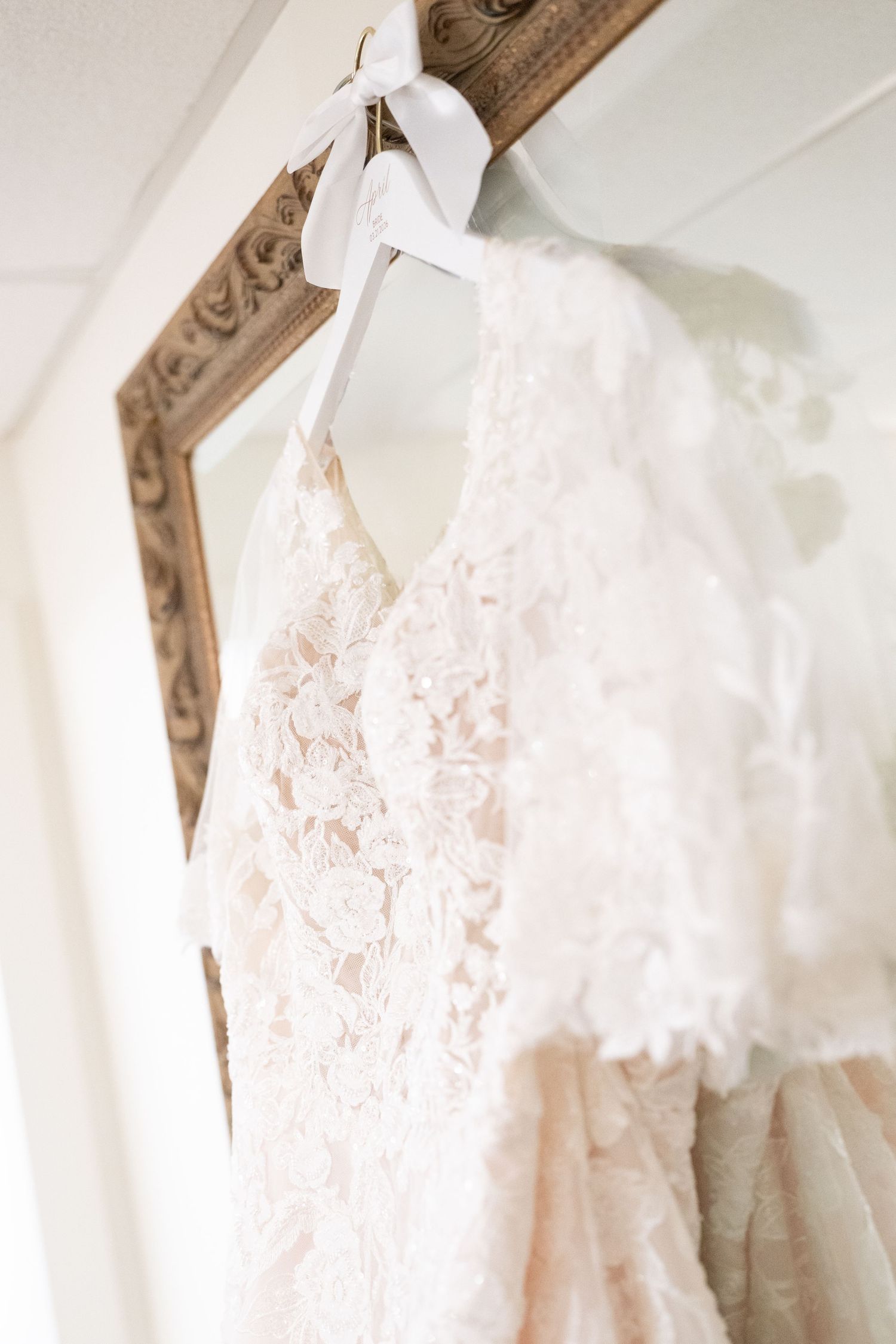 Close-up of an intricate lace bridal gown hanging on a white ribbon hanger in front of a gold mirror.