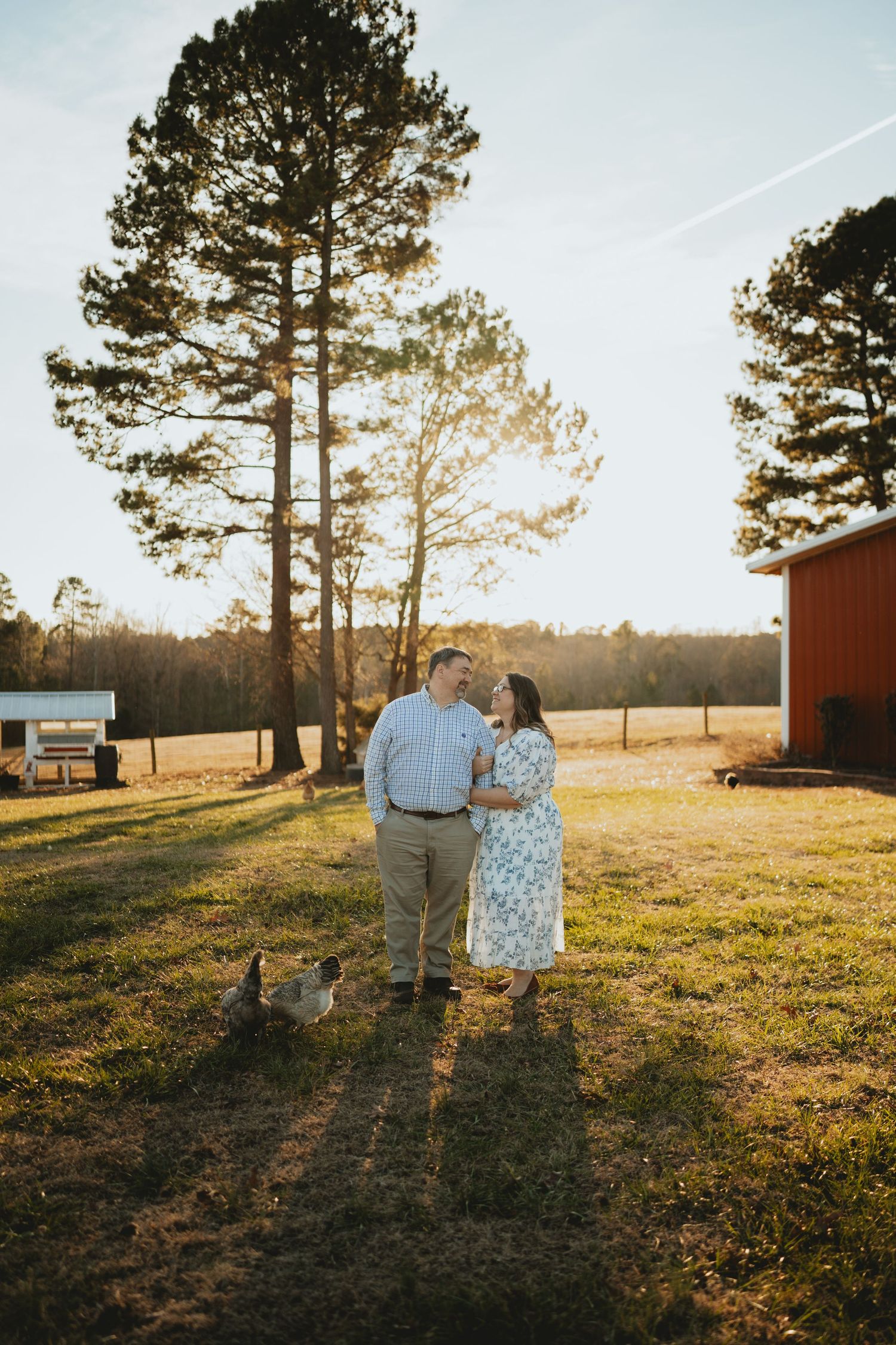 Couple holding hands and gazing at each other on a sunny farm with chickens and a red barn in the background.