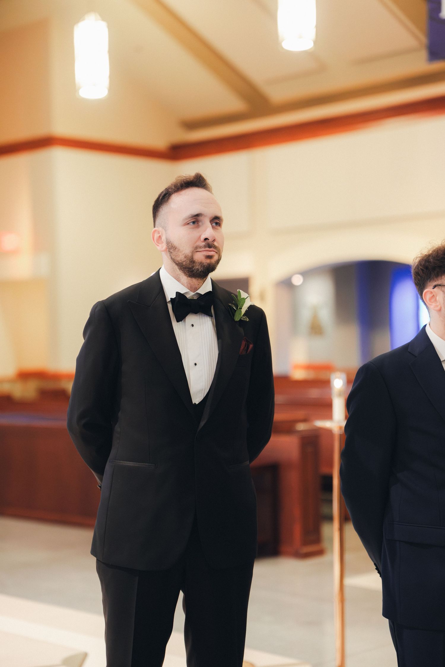 Groom in black tuxedo with boutonniere watches bride walk down church aisle.