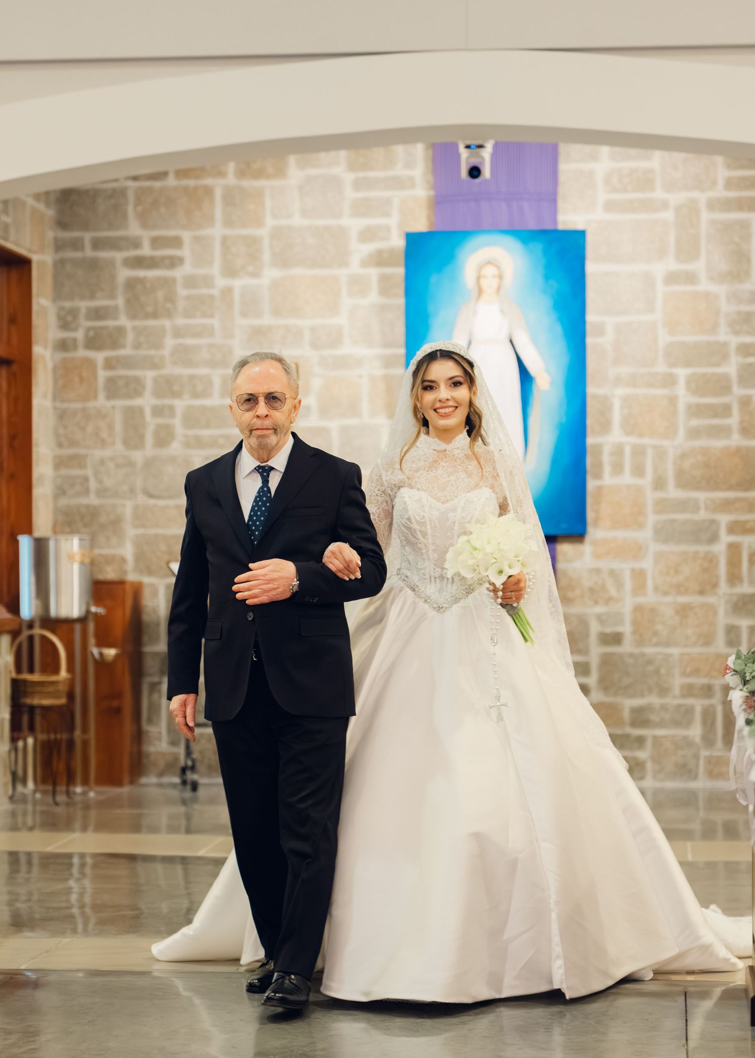 Father walking elegant bride in white ball gown down church aisle past Virgin Mary painting.