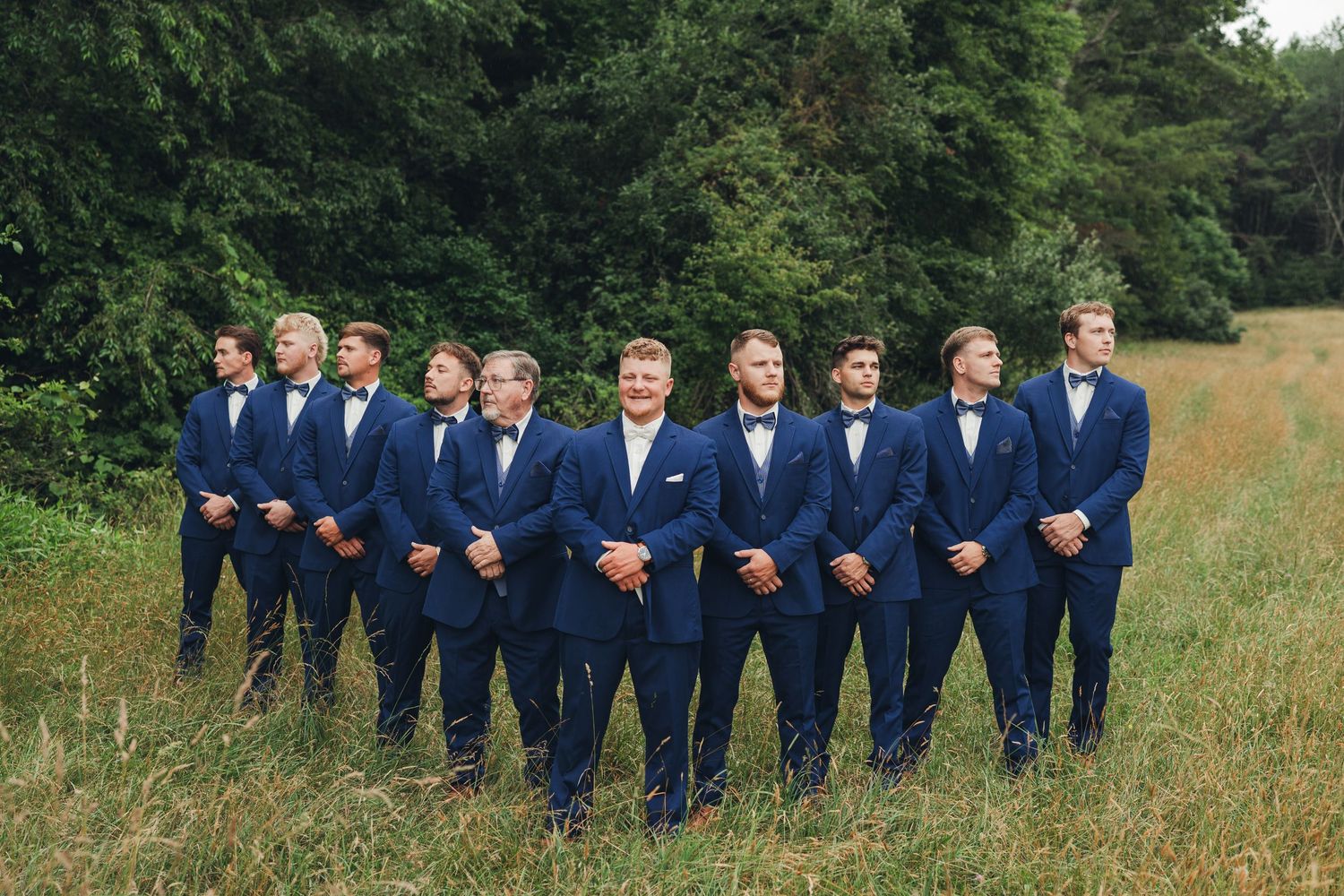 Groomsmen in navy blue suits and bow ties standing in a row outdoors in a grassy field with lush green trees.