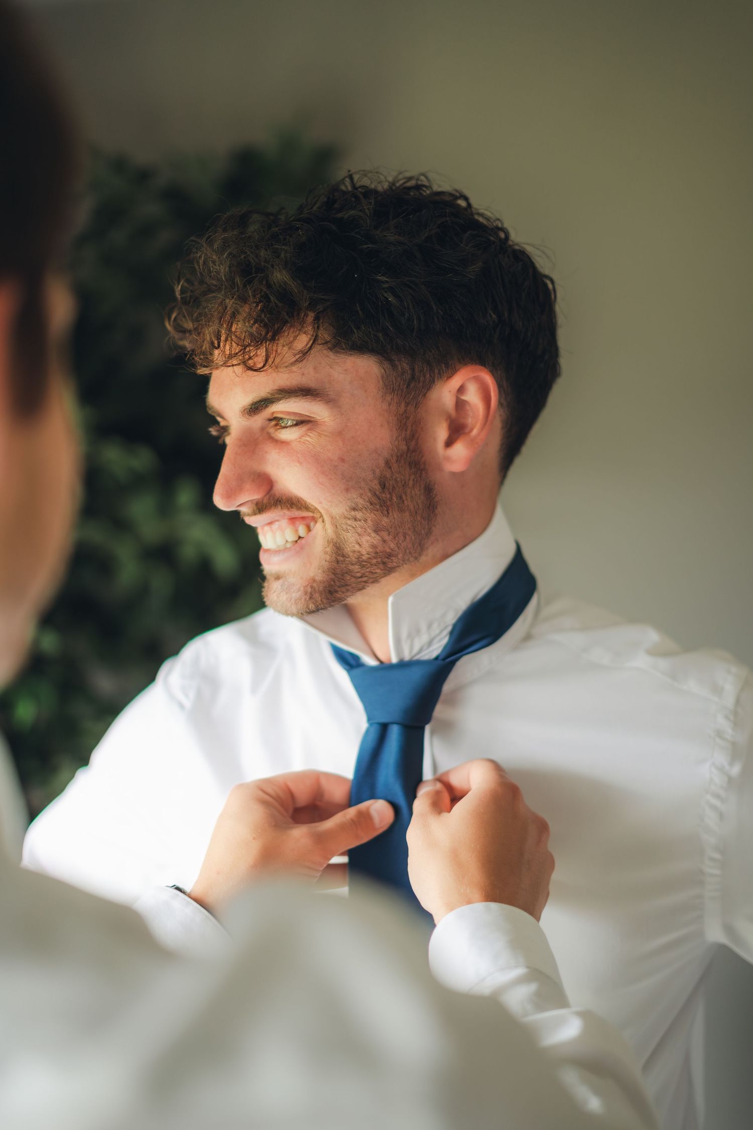 Smiling groom with curly hair getting his blue tie adjusted by a groomsman before the wedding ceremony.