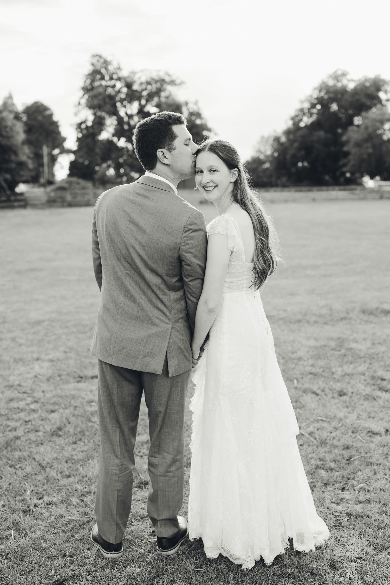 Black and white photo of groom kissing smiling bride on cheek in open field.