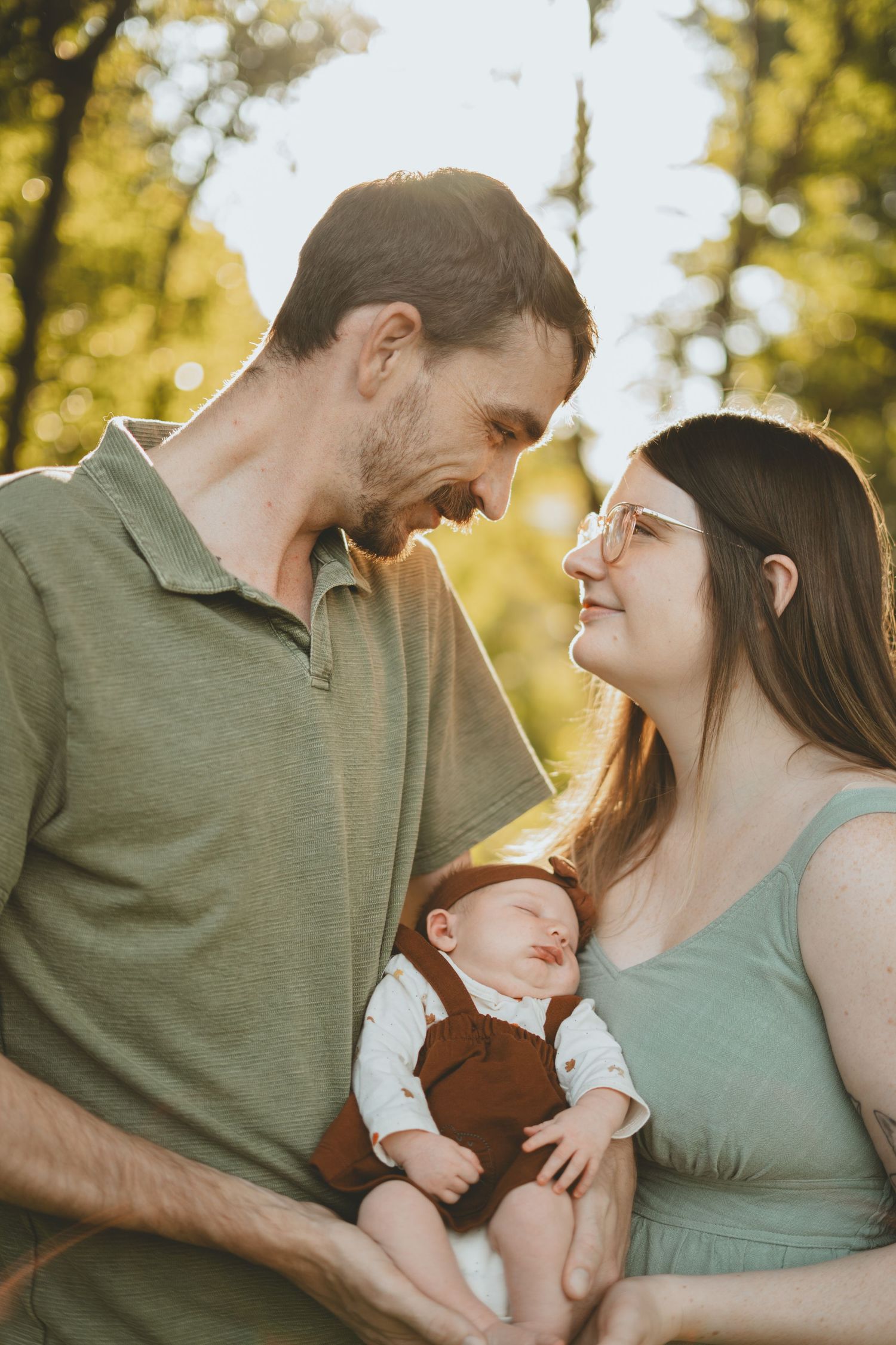 Smiling parents gaze at each other while holding their sleeping newborn baby in a sunlit outdoor setting.