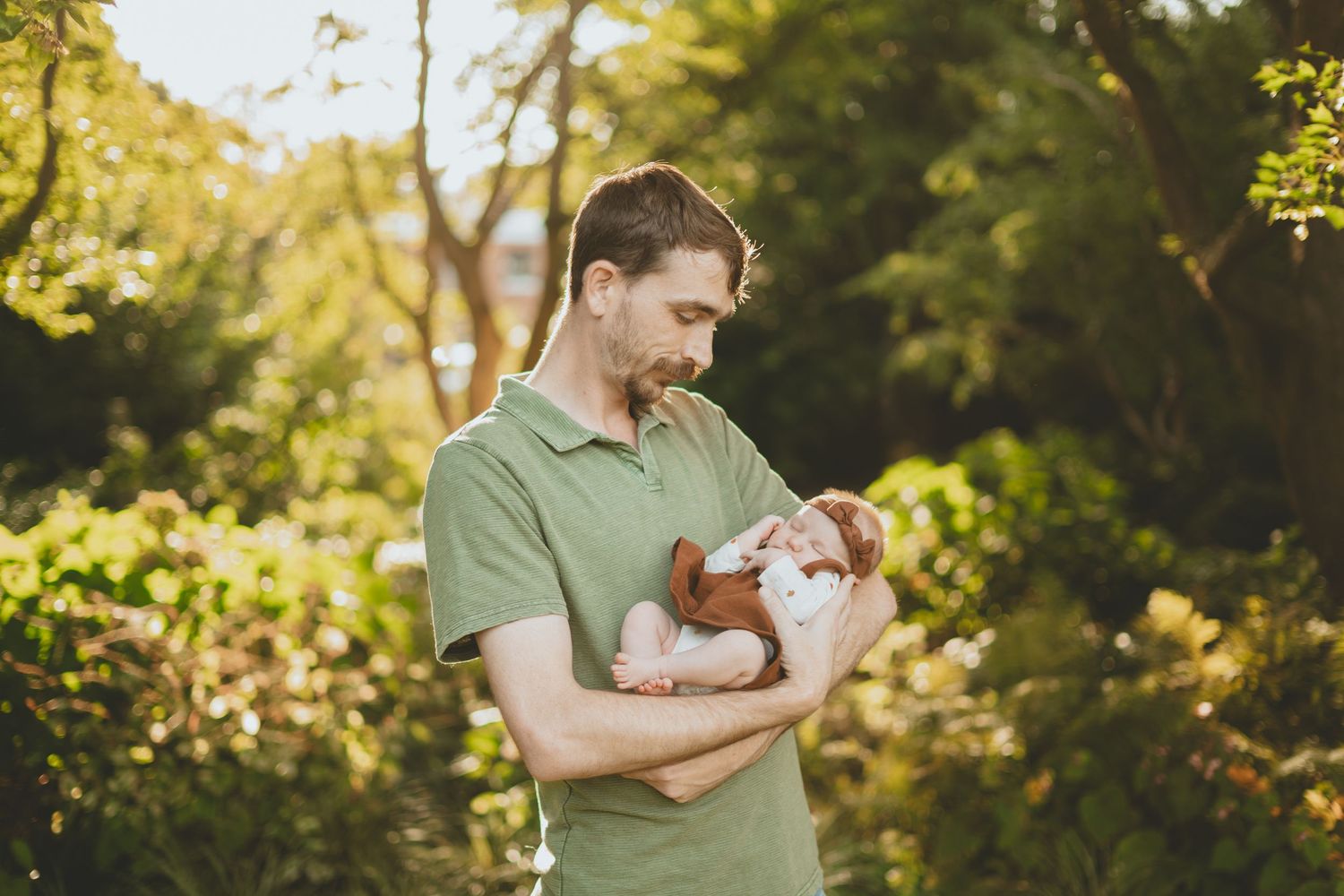 A father in a green polo shirt gently holds his sleeping newborn baby outdoors in warm sunlight.