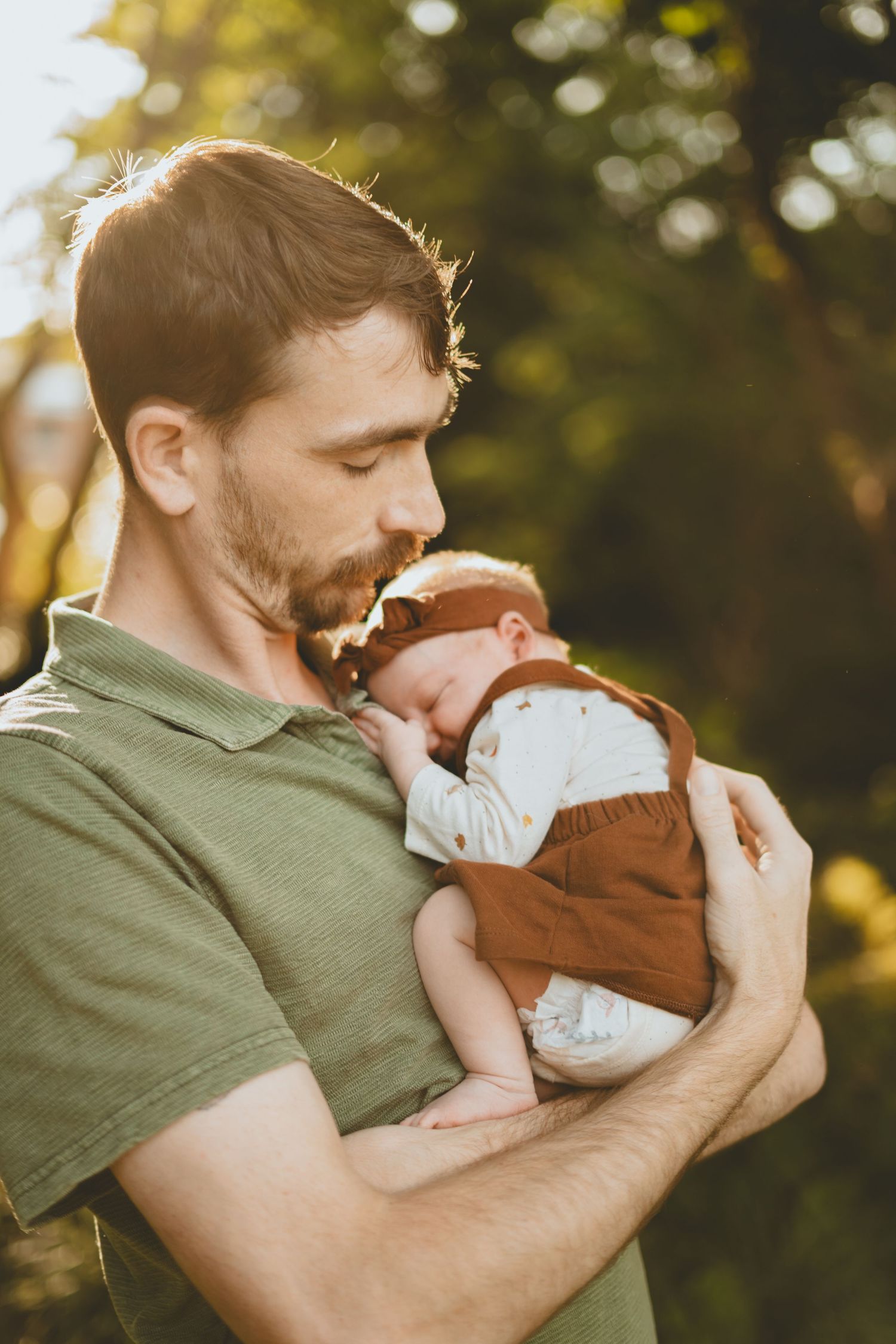 Father tenderly holds sleeping newborn baby dressed in brown outfit in warm outdoor sunlight.