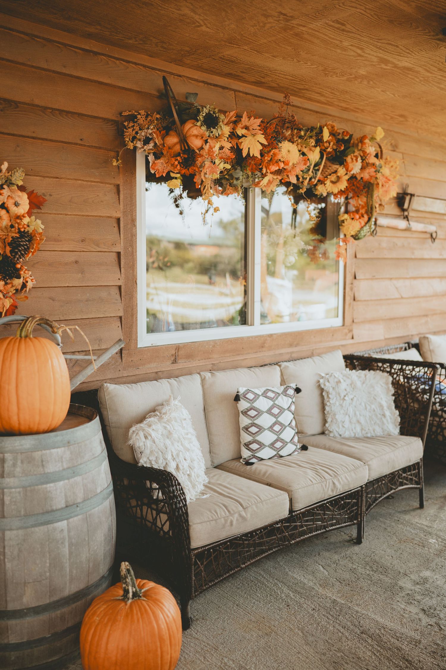 Cozy autumn porch with wicker sofa, pumpkins on barrel, and sunflower garland over window.