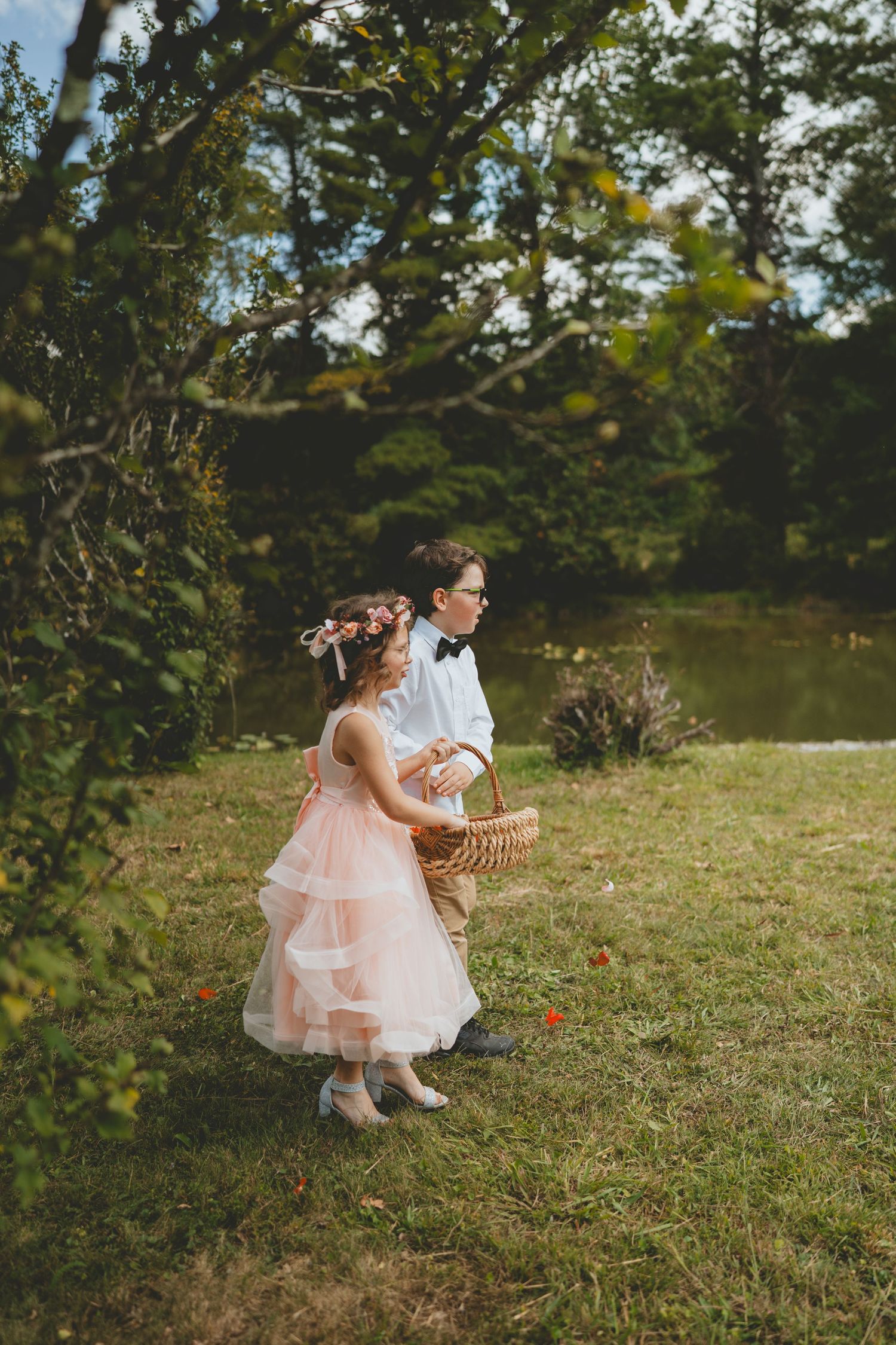 Flower girl in pink tulle dress and ring bearer carrying basket walk together near a pond.