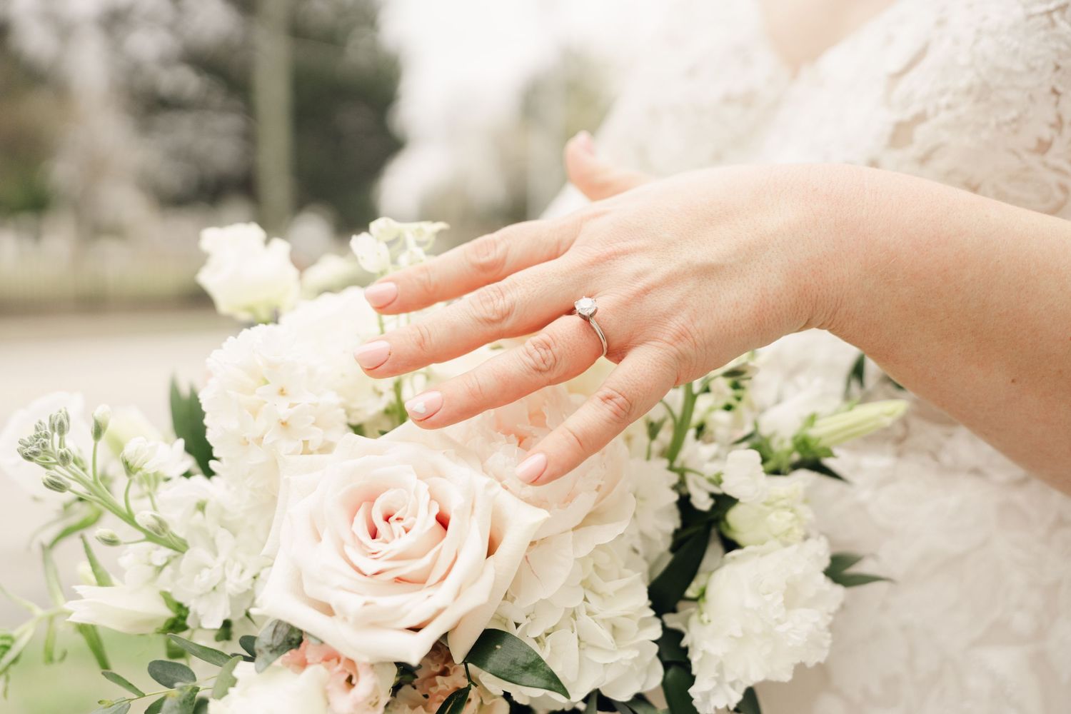 Close-up of bride\u2019s hand with diamond ring resting on white and blush rose wedding bouquet.