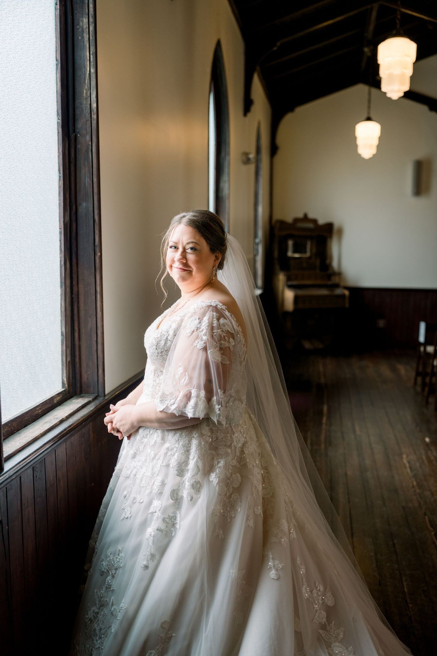 Elegant bride in lace wedding gown and veil standing by window in historic church.