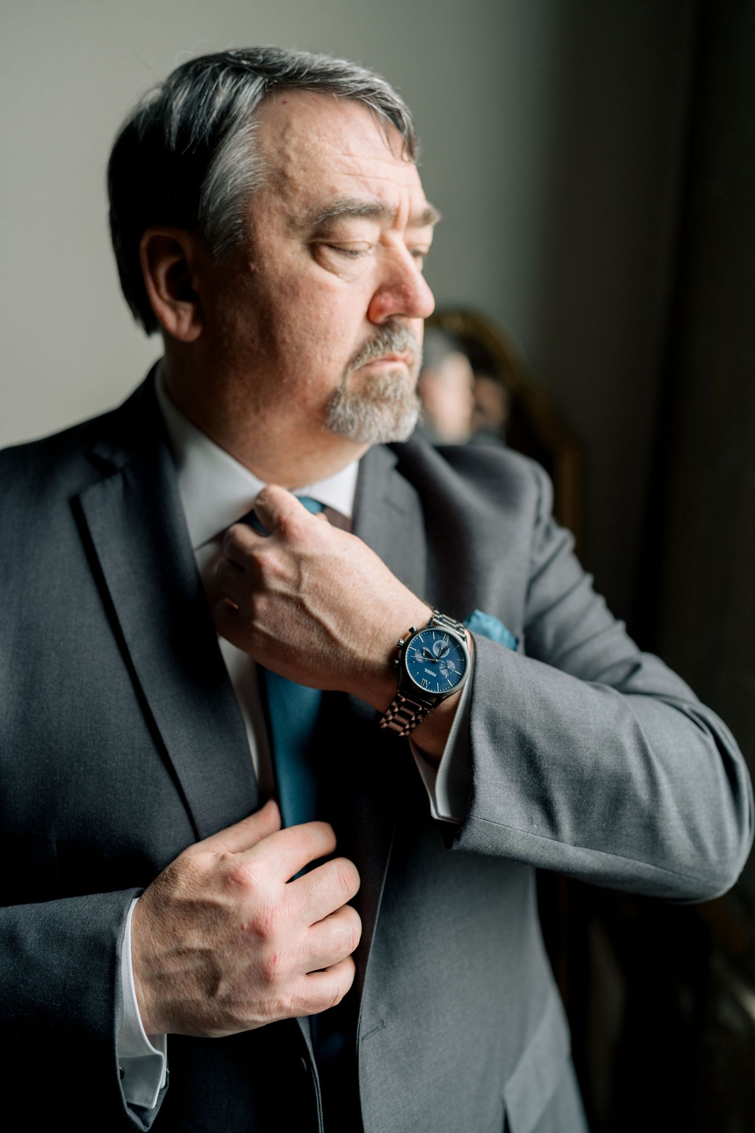Distinguished groom in gray suit adjusting his tie while wearing a classic blue chronograph watch before the wedding.