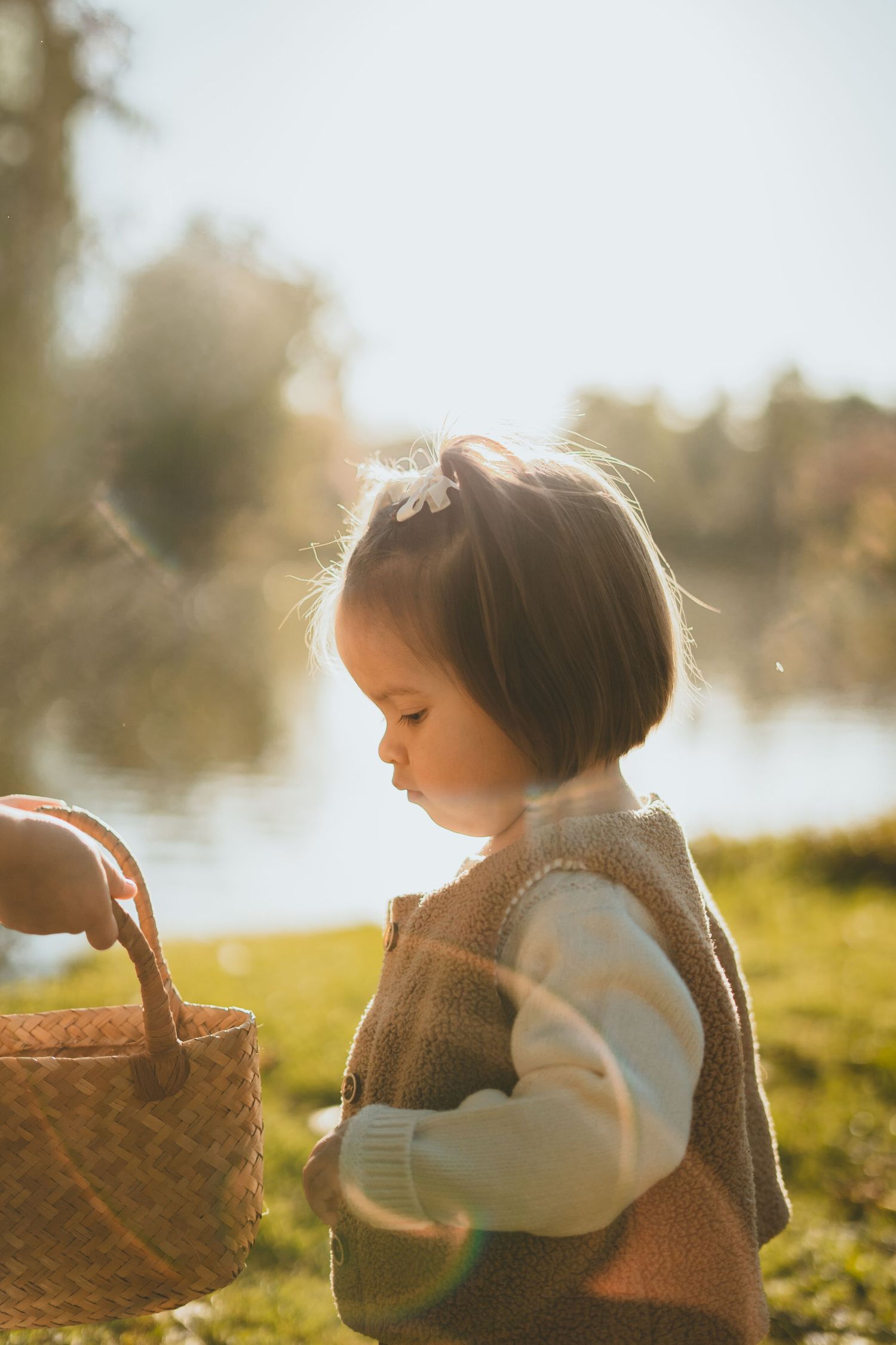 Toddler girl in knit vest holding a wicker basket by a sunny lake.
