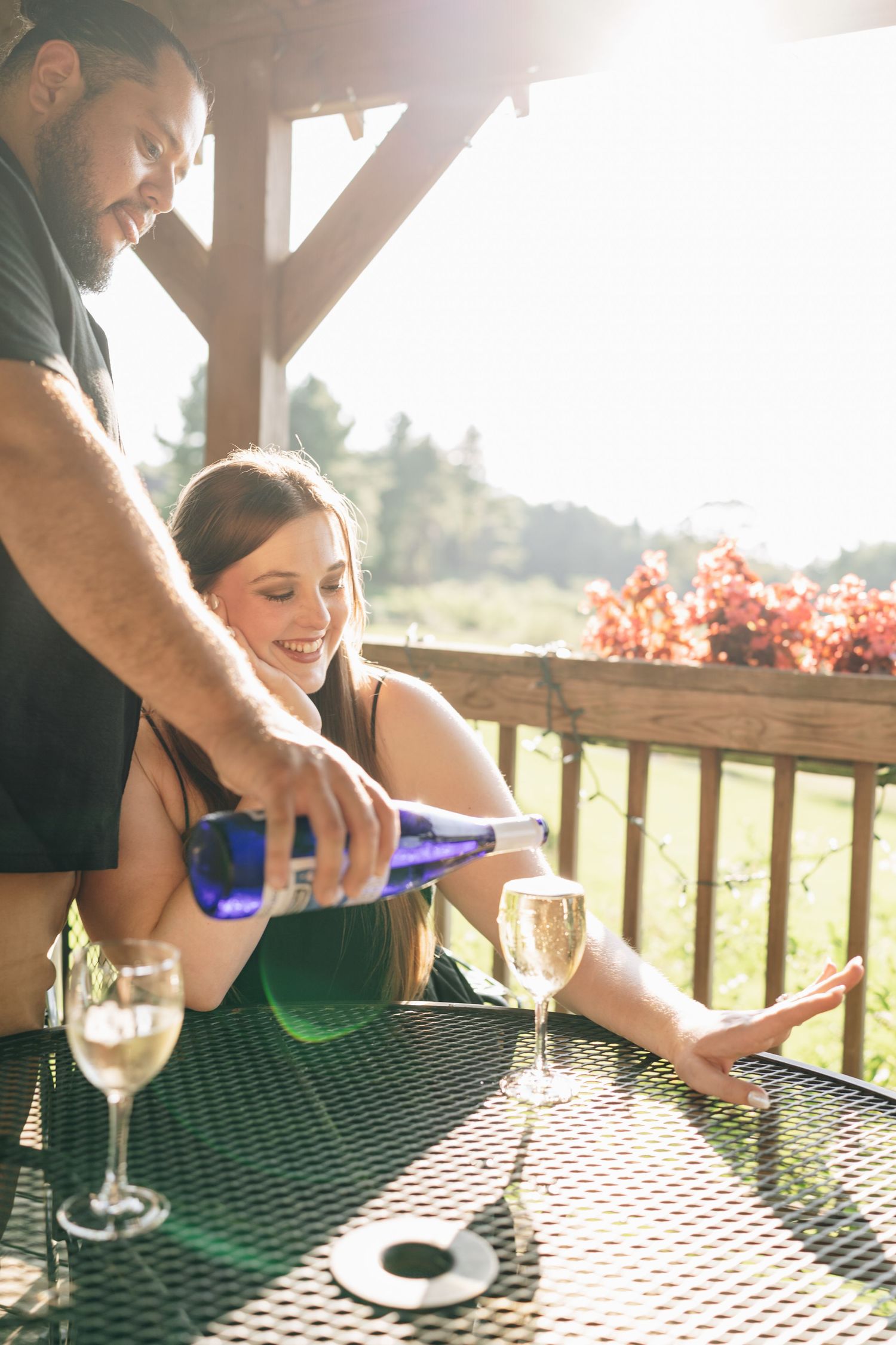 Man pouring white wine from blue bottle for smiling woman on sunny outdoor patio with garden views.