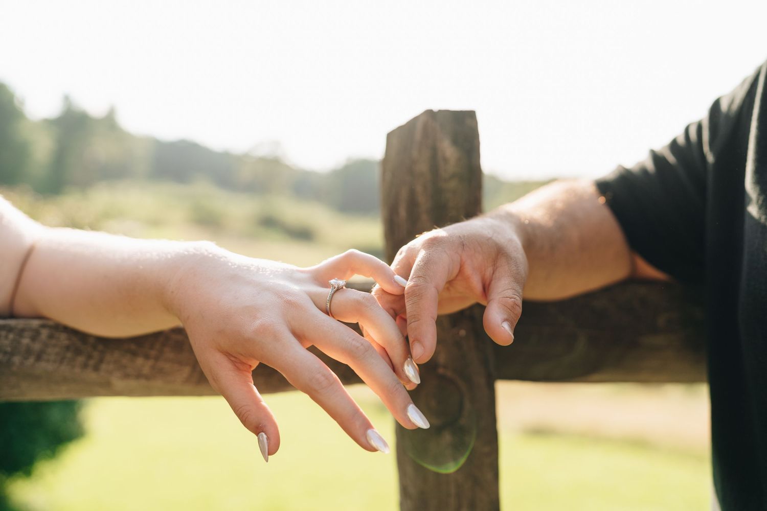 Close-up of couple exchanging rings outdoors near a wooden fence with green hills in the background.