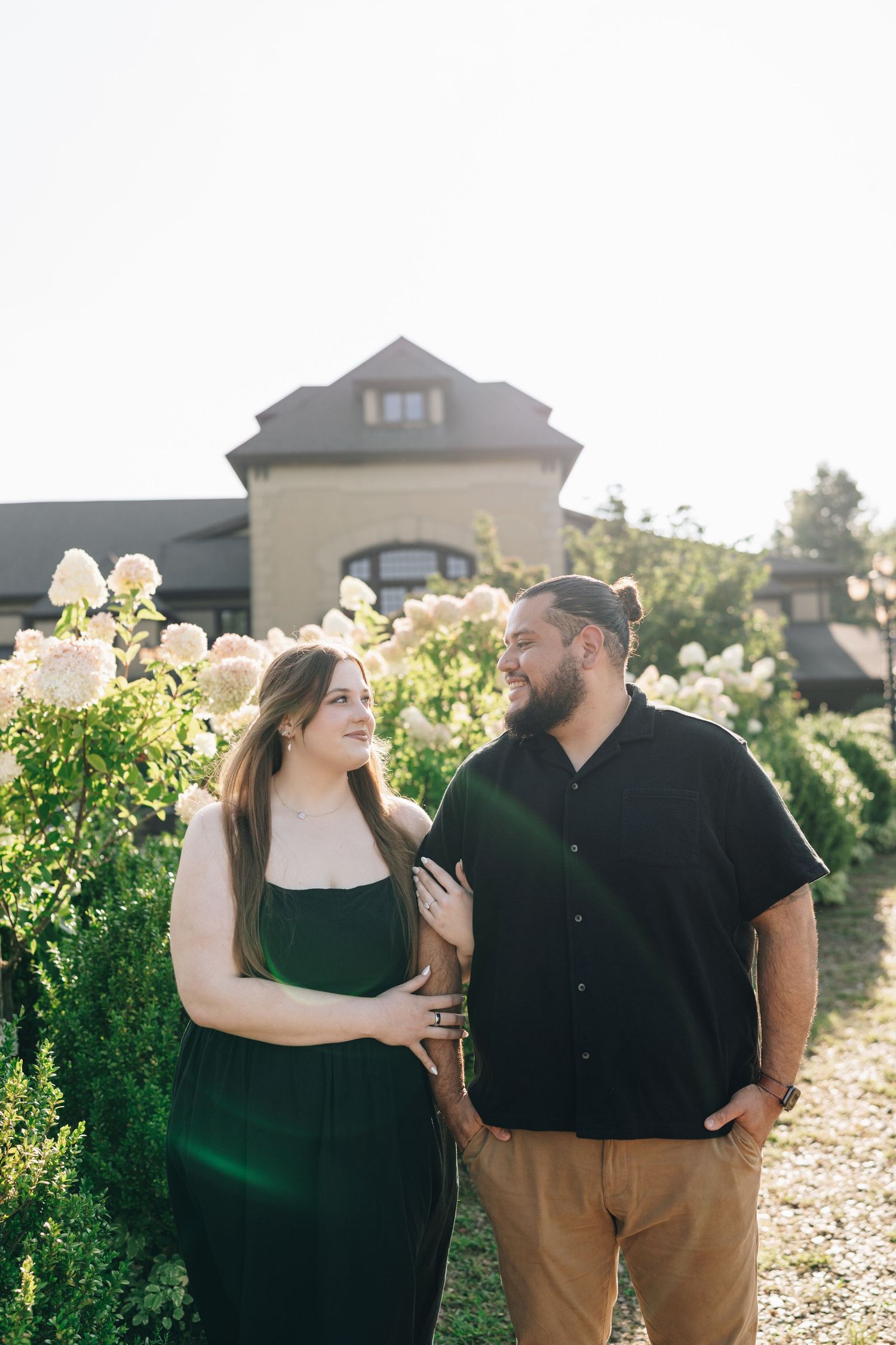 Engaged couple smiling at each other among white hydrangeas outside an elegant estate building.