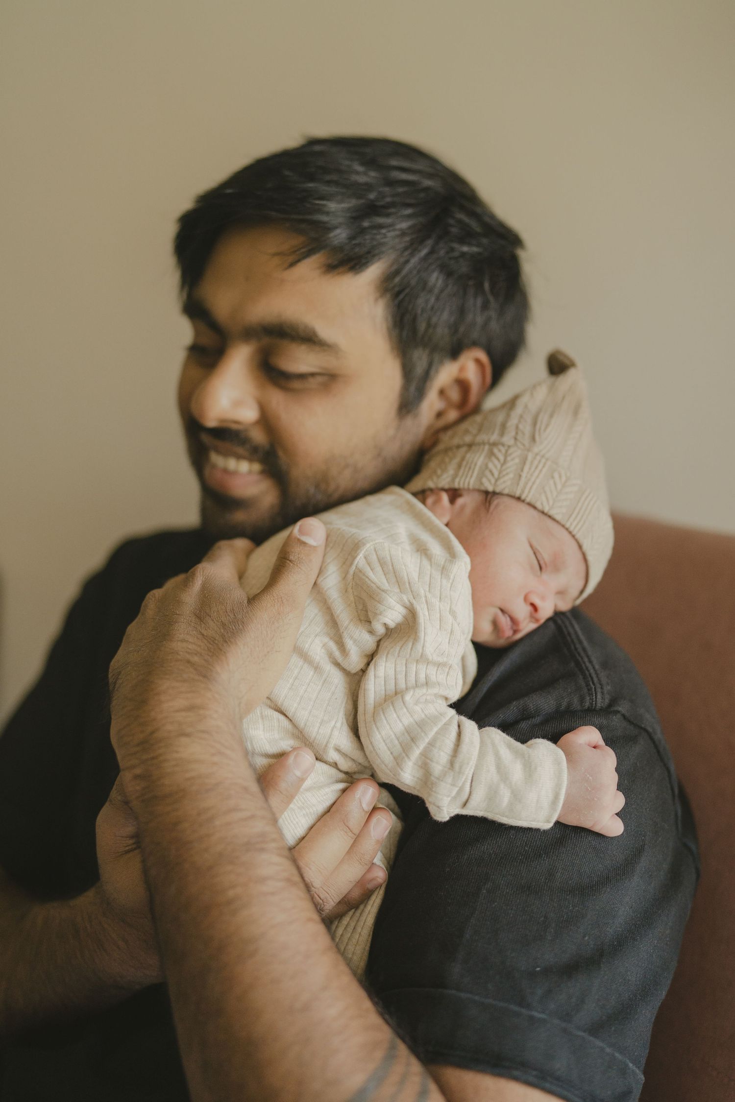 Bearded father smiling warmly while holding his sleeping newborn baby in a knit hat.