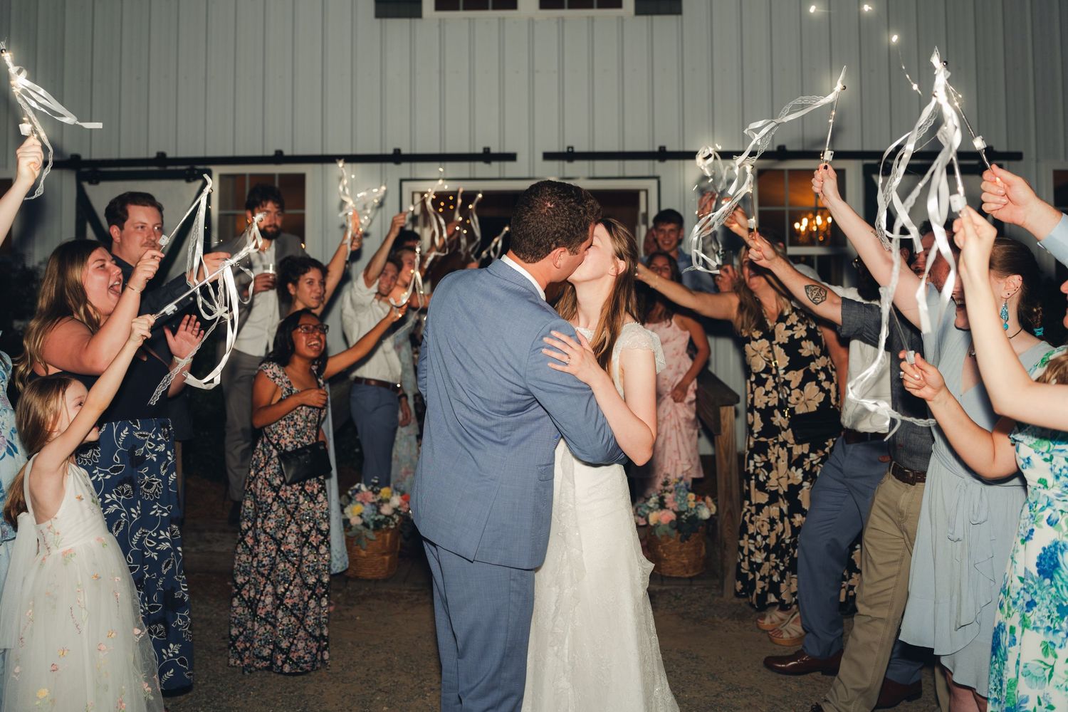 Bride and groom share a kiss during a ribbon wand sendoff surrounded by cheering wedding guests at a barn reception.