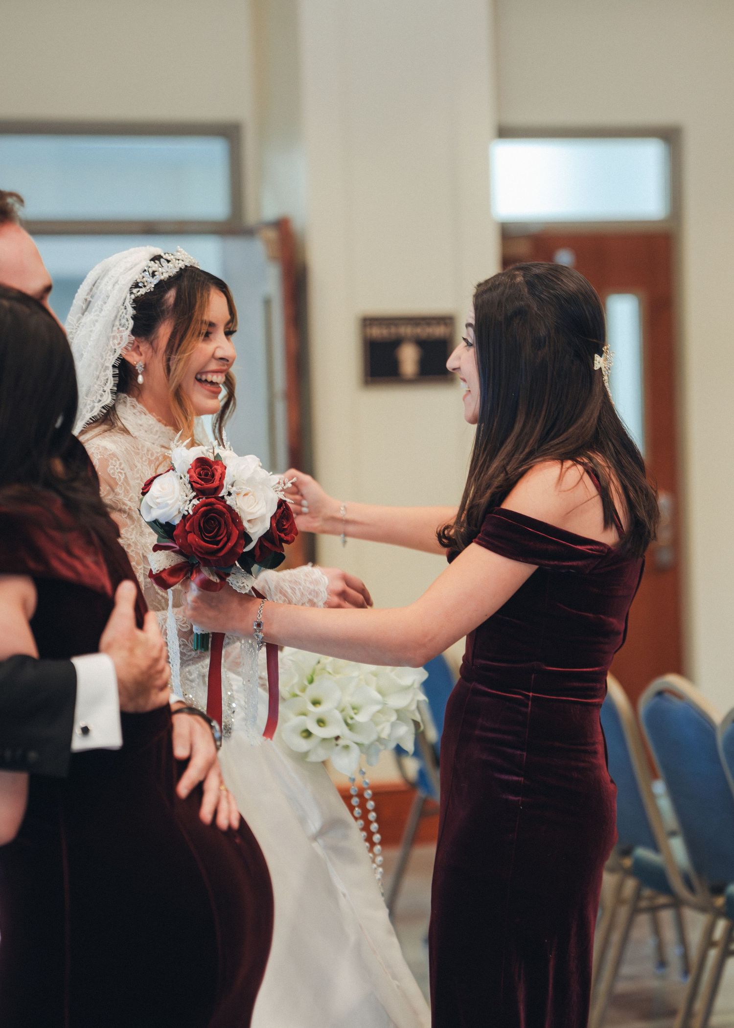 Bridesmaid in burgundy velvet dress hands bouquet to laughing bride at wedding reception.