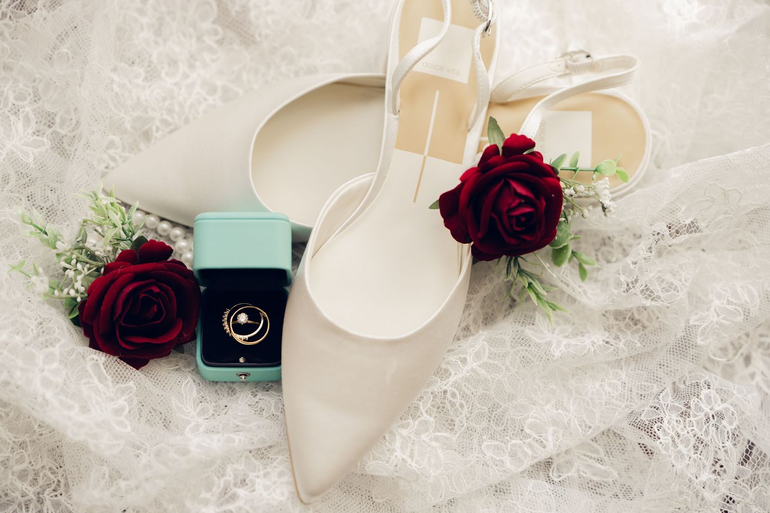 Bridal accessories flat lay with ivory heels, red roses, and a gold ring in a teal box.