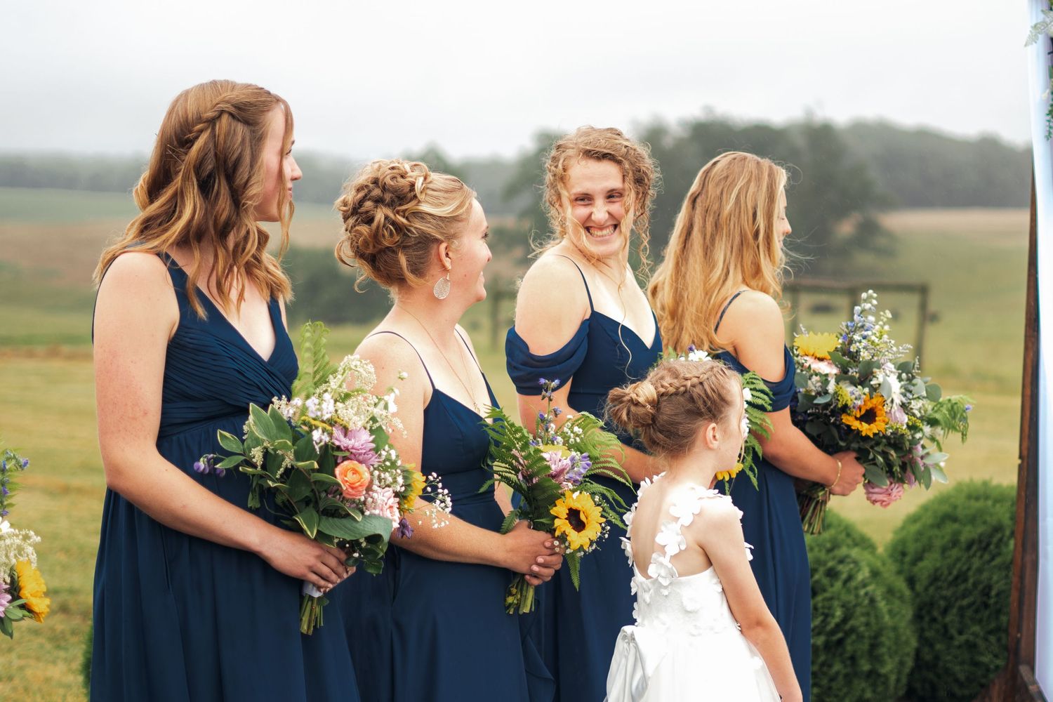 Bridesmaids in navy blue dresses holding sunflower bouquets with a flower girl in white at an outdoor wedding.