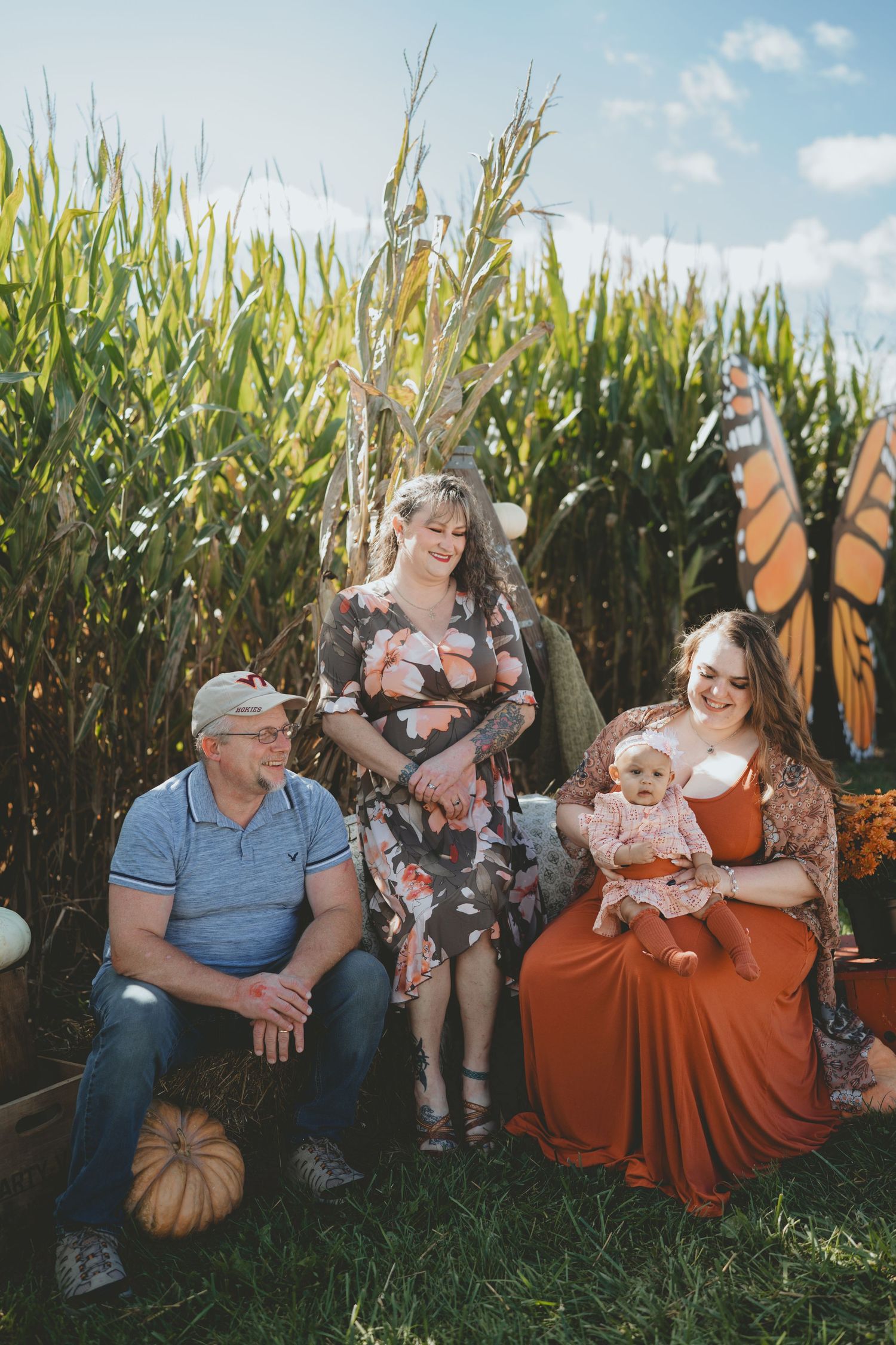 Family of four with baby sitting near cornfield with butterfly decorations and pumpkins.
