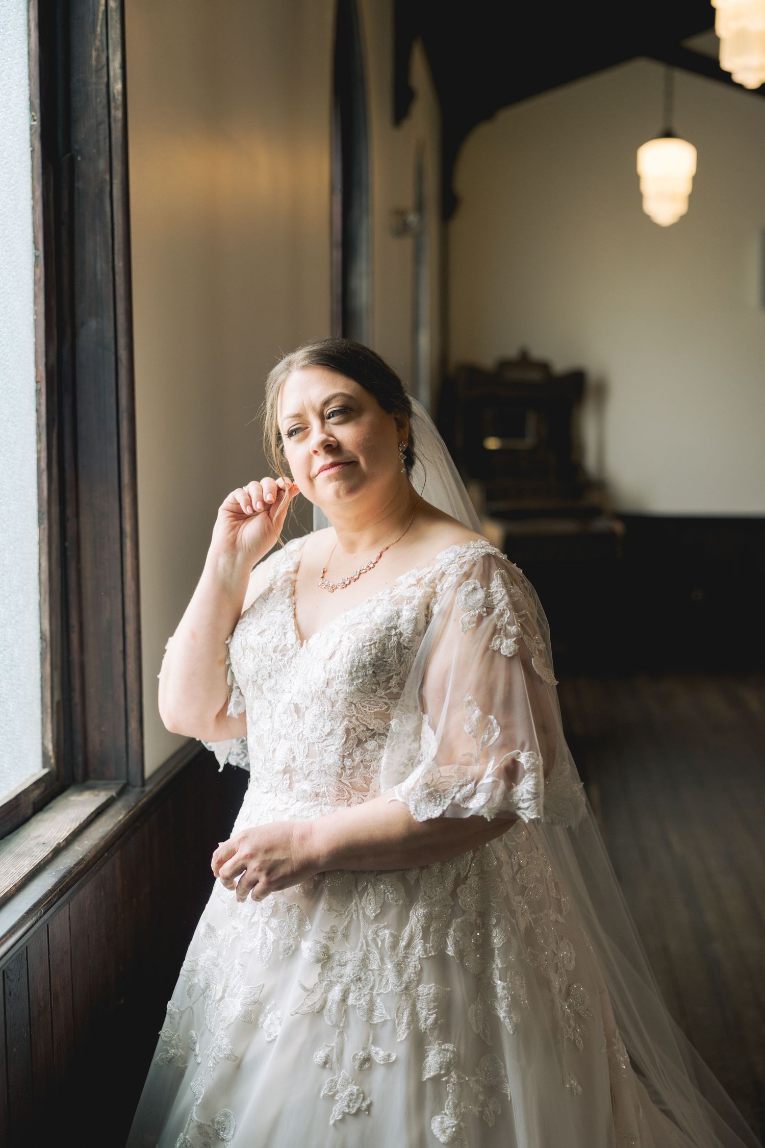 Bride in lace wedding gown and veil poses by window in elegant indoor setting.