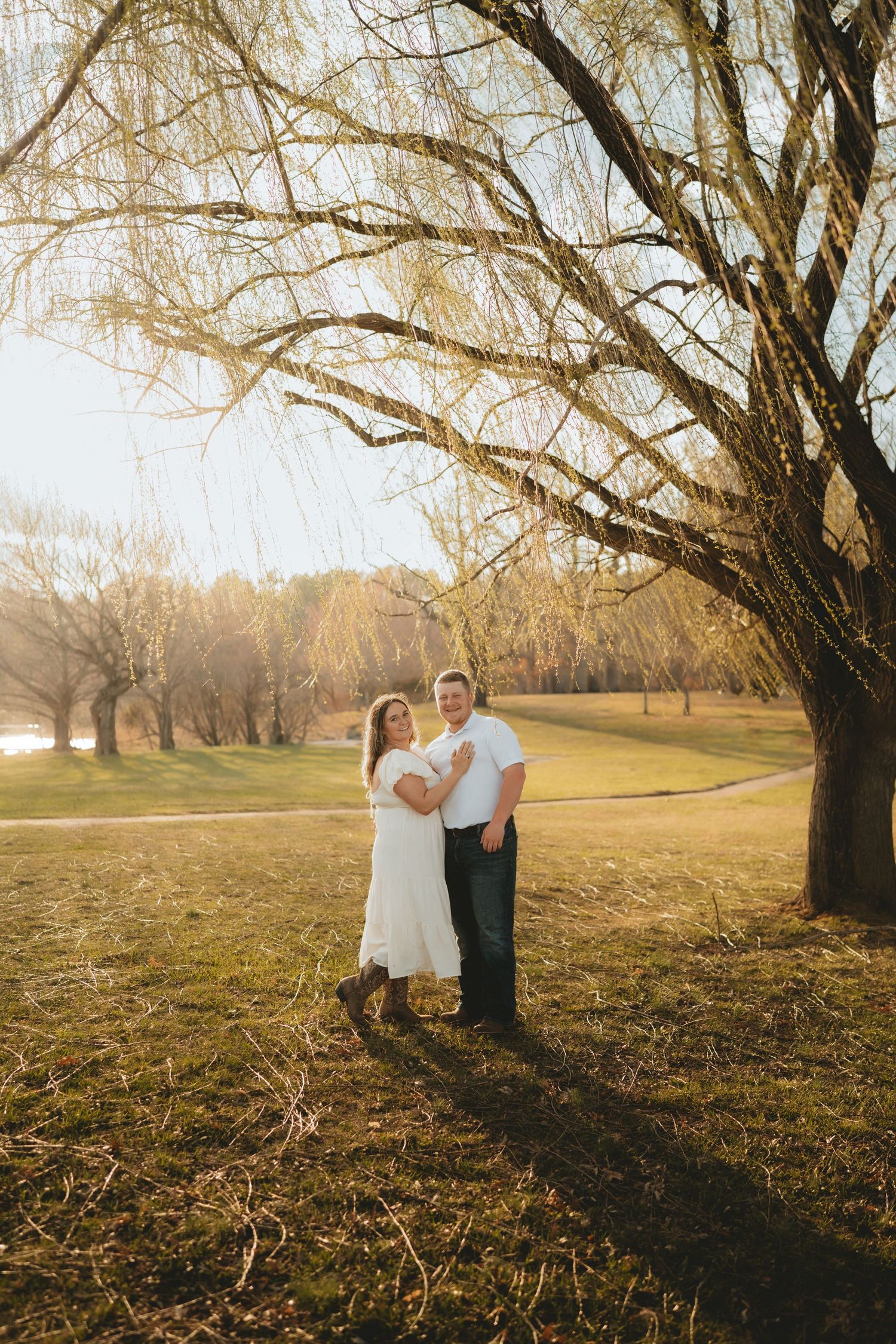 Couple embracing under a willow tree in golden hour sunlight during an outdoor engagement session.