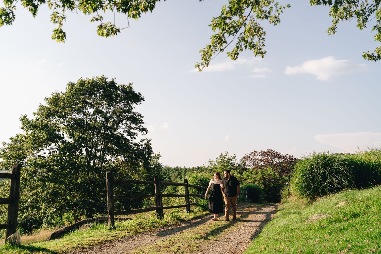 Couple walking hand-in-hand down a sunlit gravel path surrounded by lush green trees and meadows.