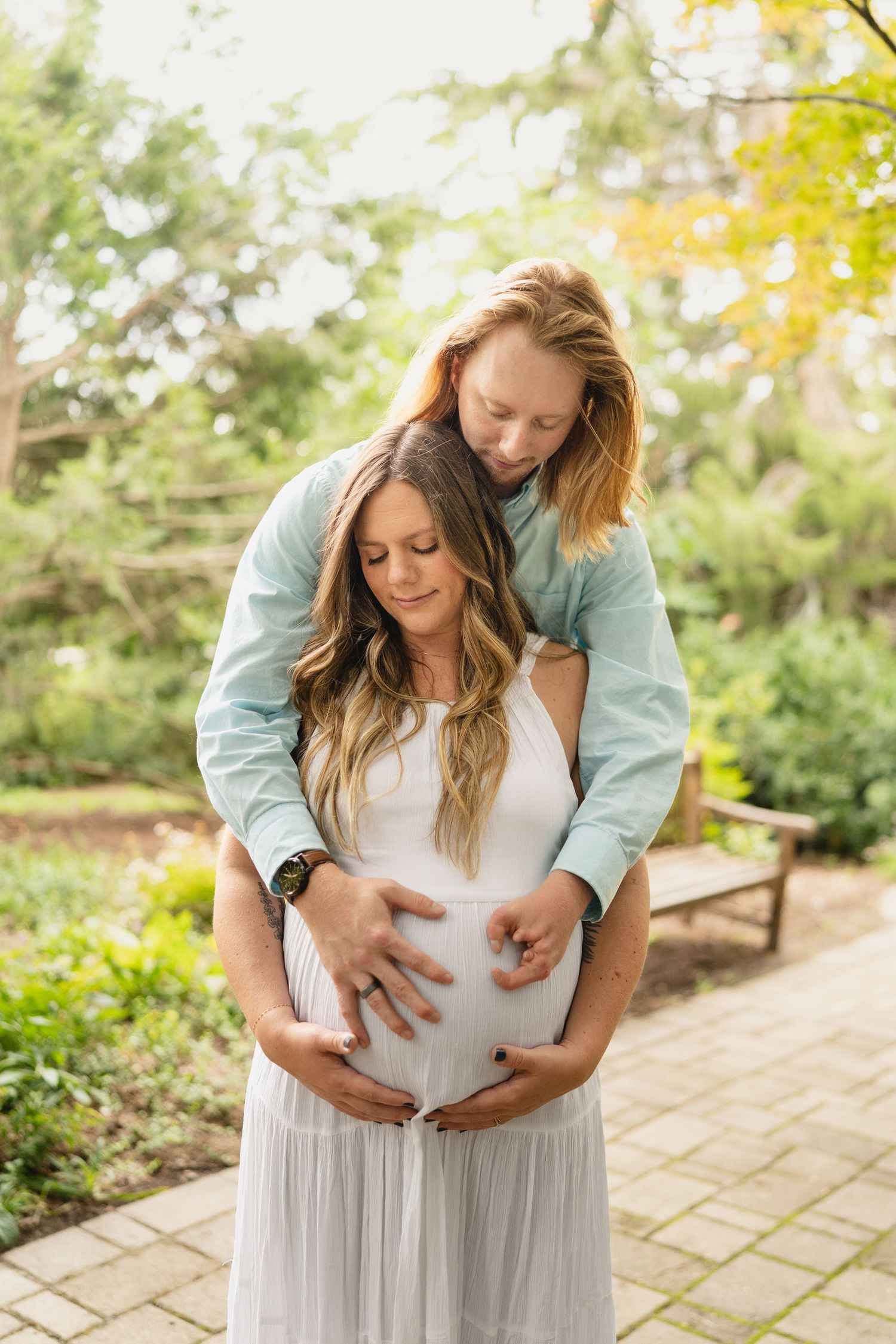 Couple tenderly cradles pregnant belly outdoors in lush green garden setting.