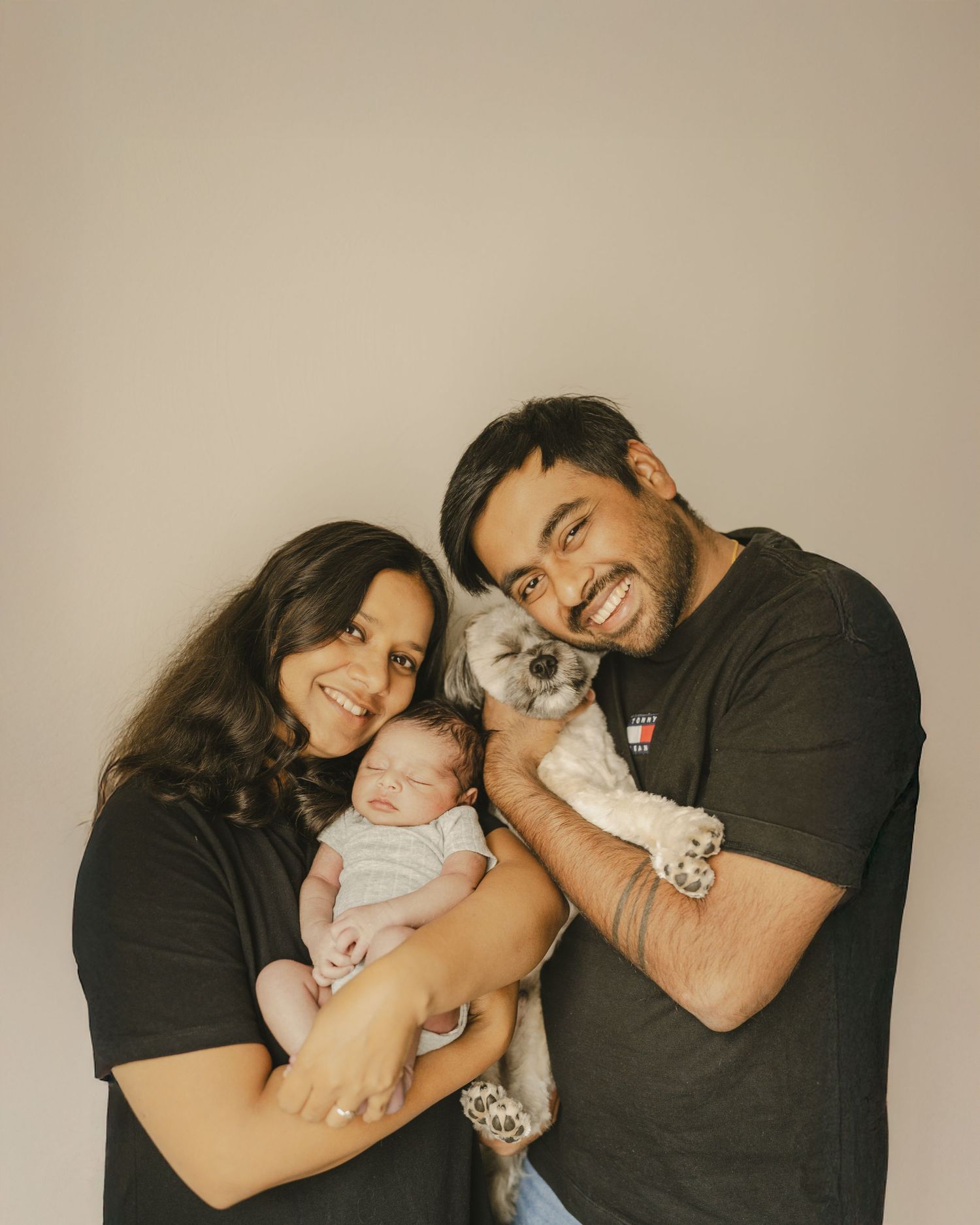 Happy couple in black shirts holding newborn baby and small fluffy dog in studio portrait.