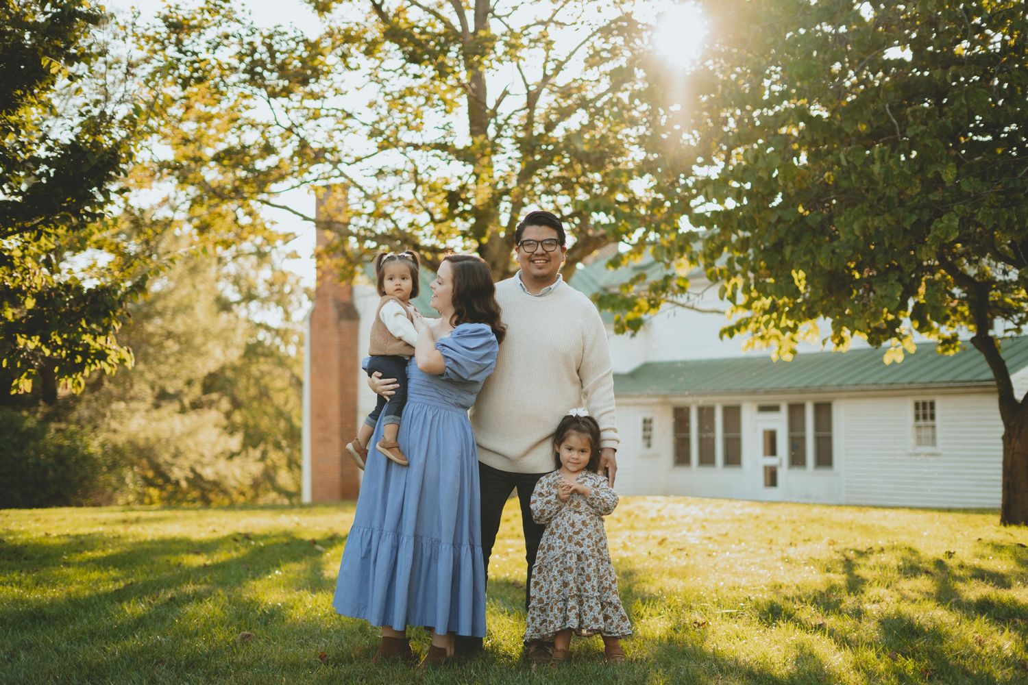 Happy family with two young daughters posing outdoors in golden sunlight near a white farmhouse in autumn.
