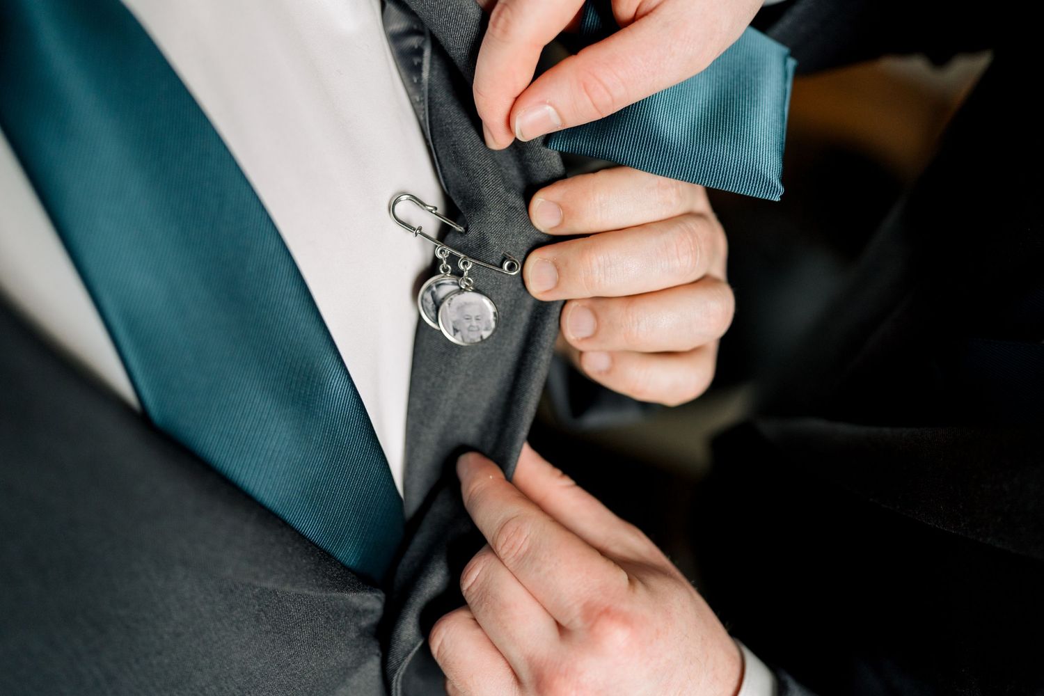 Close-up of groom's boutonniere with heart charm being pinned to dark teal suit lapel.