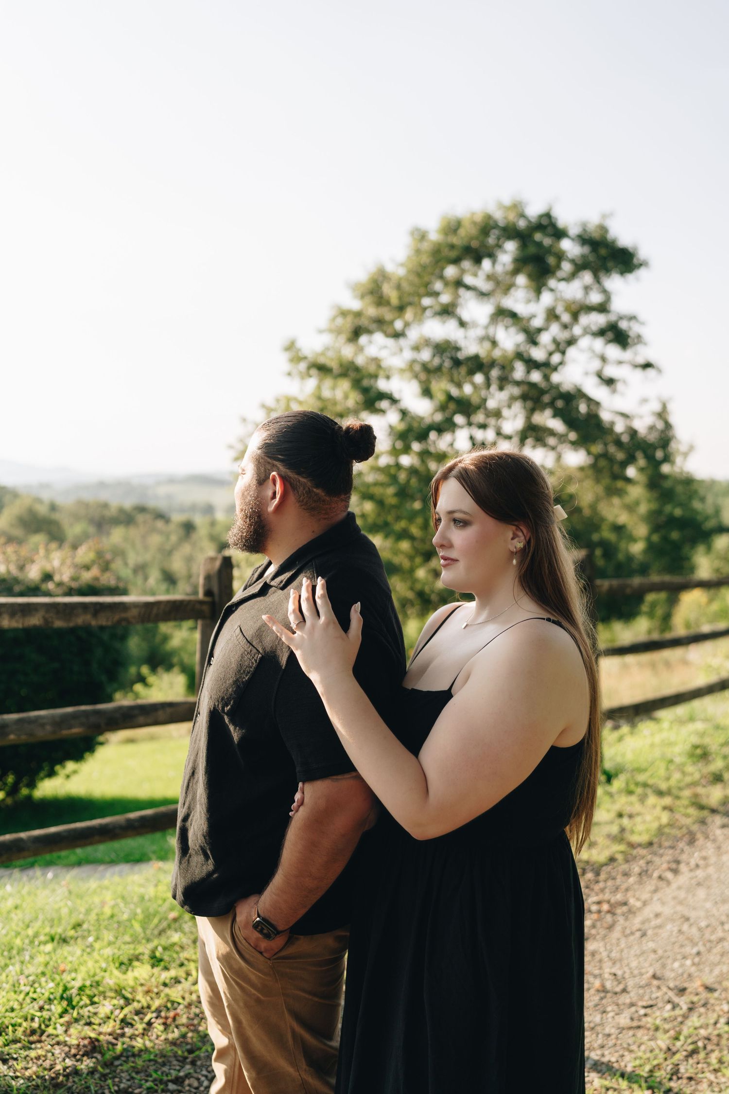 Couple in black outfits embracing near a rustic wooden fence with scenic green landscape behind them.