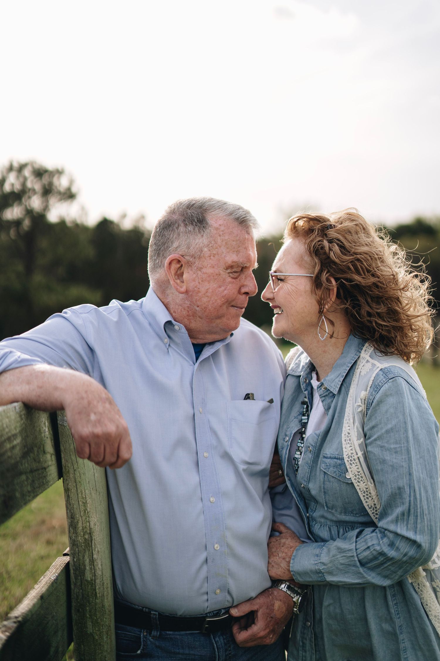 Older couple sharing a tender moment leaning on wooden fence, smiling at each other outdoors.
